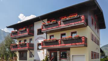 Ein mehrstöckiges Gebäude mit Balkonen, die mit roten Blumen geschmückt sind, einer steinernen Fassade und einem wolkenlosen Himmel im Hintergrund.