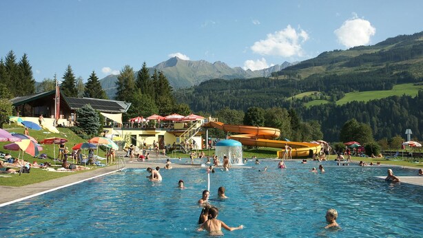Ein Schwimmbad mit Badegästen, einer Wasserrutsche und einer Bergkulisse.