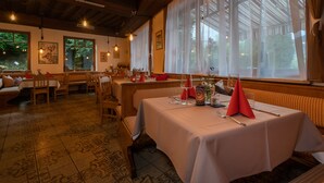 Ein Restaurant mit Holztischen und -stühlen, großen Fenstern mit Vorhängen und hängenden Lampen.