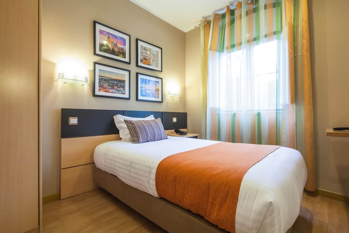 A neatly made bed with a white and orange blanket, a wooden headboard, and a nightstand with a phone. There are framed pictures on the wall and a window with striped curtains.