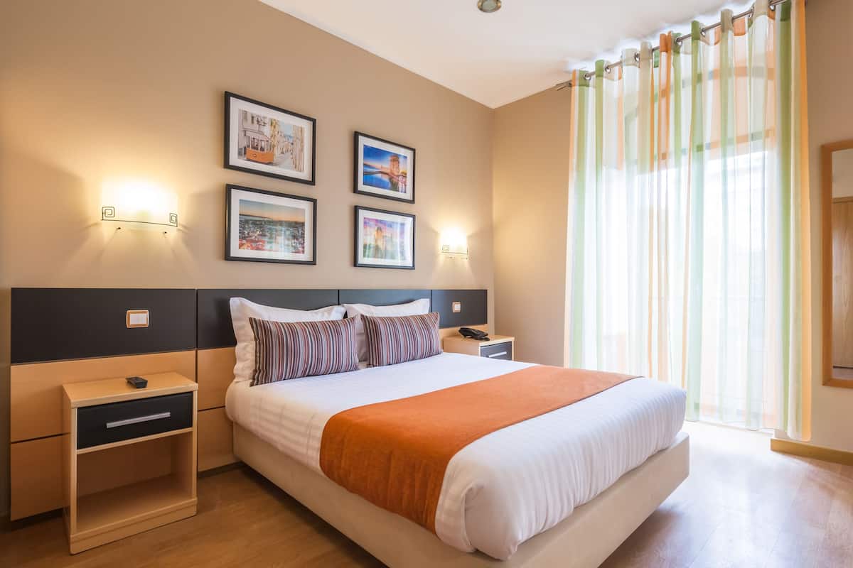 A hotel room with a large bed, two bedside tables, a nightstand, and a window with sheer curtains.