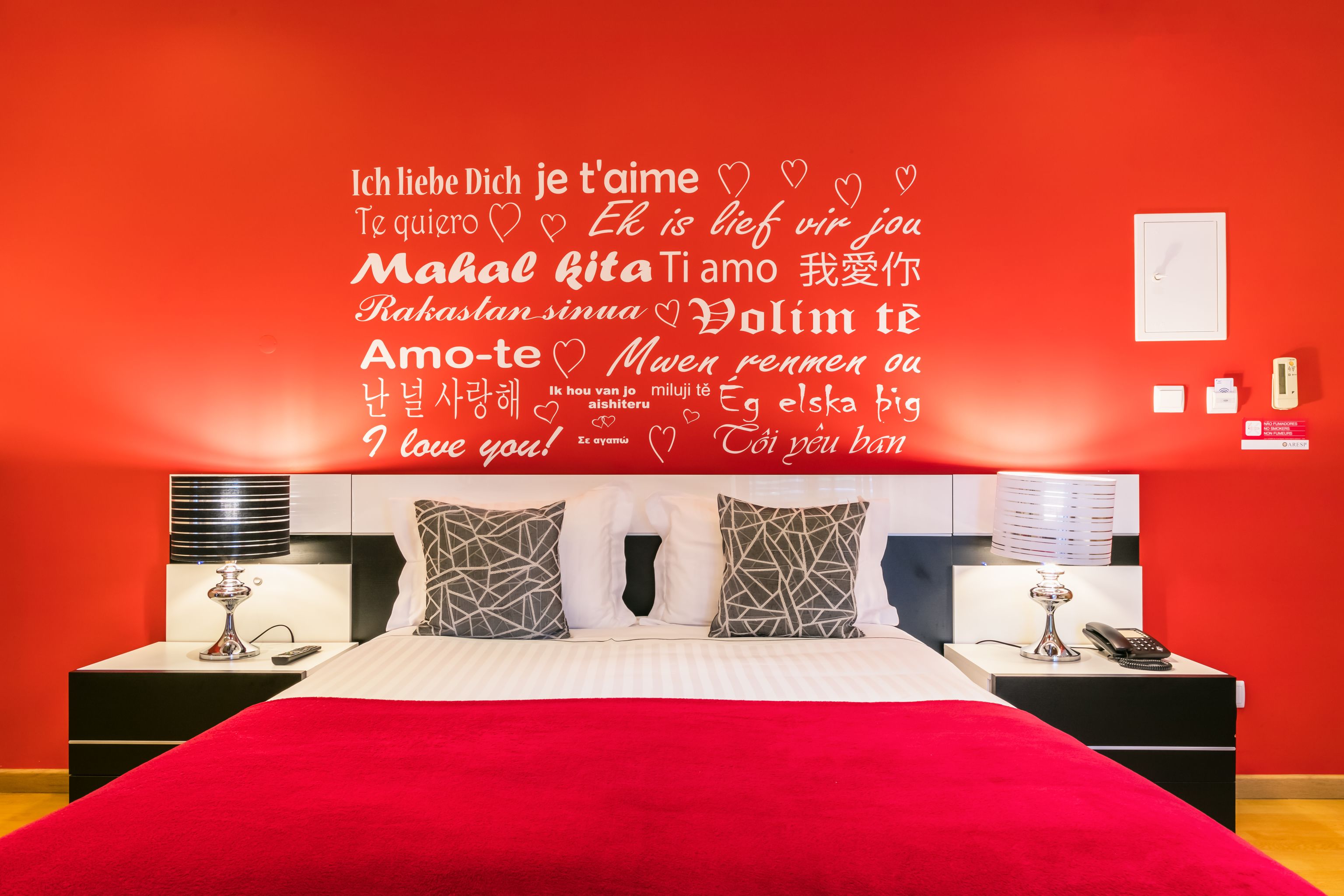 Habitación de hotel con pared roja, cama con manta roja y pared decorada con citas de amor en varios idiomas.