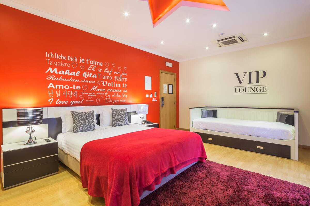 A hotel room with a large bed, a smaller bed, a red wall with multilingual love quotes, and a sign reading "VIP LOUNGE".