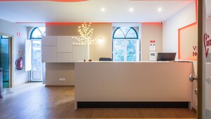 A reception area with a modern design, featuring a reception desk, a decorative light fixture, and a view through the windows. - Vila Nova Guesthouse (Lisbon)