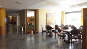 Comedor de hotel con una mesa redonda y sillas, plantas en macetas y un ventanal con cortinas.