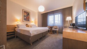 Ein Hotelzimmer mit einem Bett, Nachttischlampen, einem Schreibtisch mit Fernseher, einem Sessel und einem Fenster mit Vorhängen.