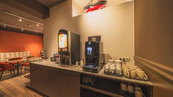 Una zona de bar con dos máquinas de café, un adorno con forma de barco y un espacio de estar con sillas rojas y una mesa de madera.