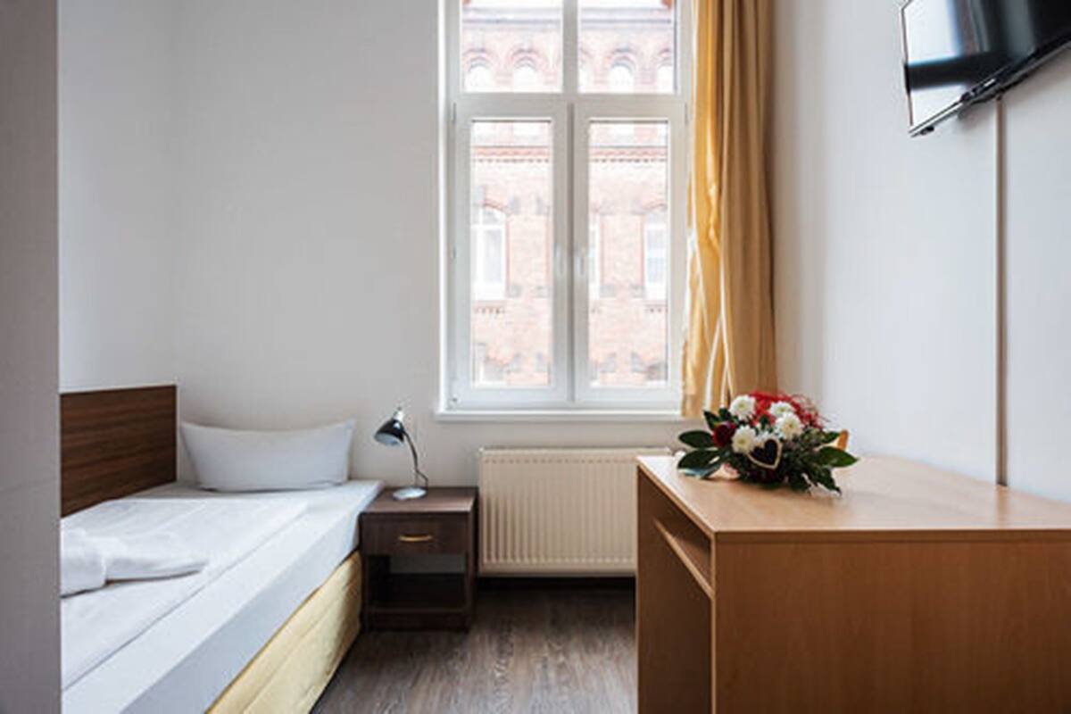 Una habitación de hotel con cama, mesita de noche, lámpara, escritorio y televisor en la pared.