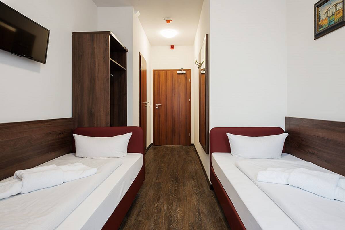 Habitación de hotel con dos camas individuales, un armario de madera, un televisor en la pared y una puerta que da a otra habitación.