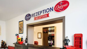 A reception area with a sign reading "RECEPTION" and "Queen's Garden". There are red chairs, a telephone booth, and a shelf with binders. - Hotel Queenspark (Berlin)