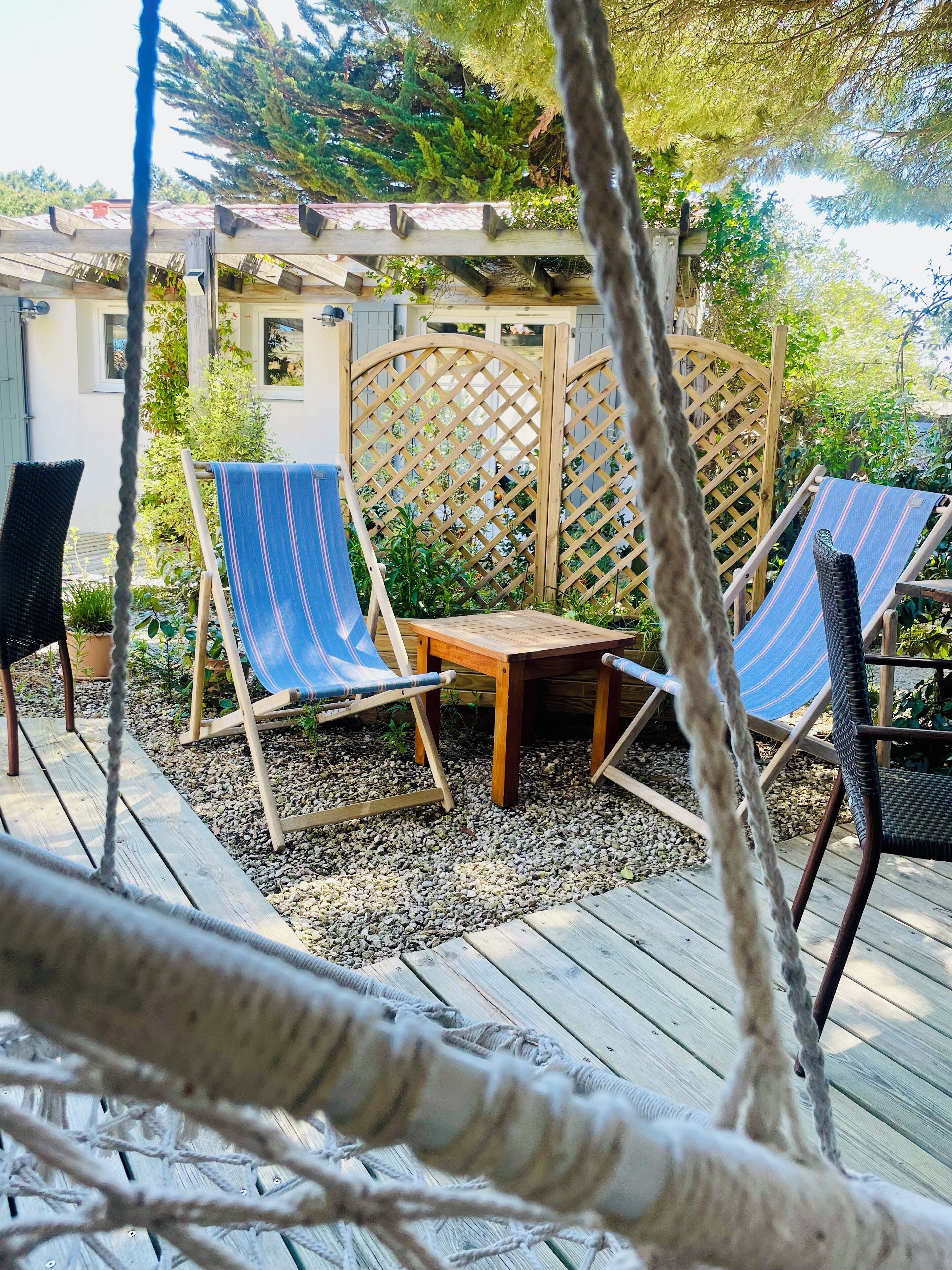 A wooden deck with two striped lounge chairs and a small table, surrounded by a wooden lattice fence and greenery.