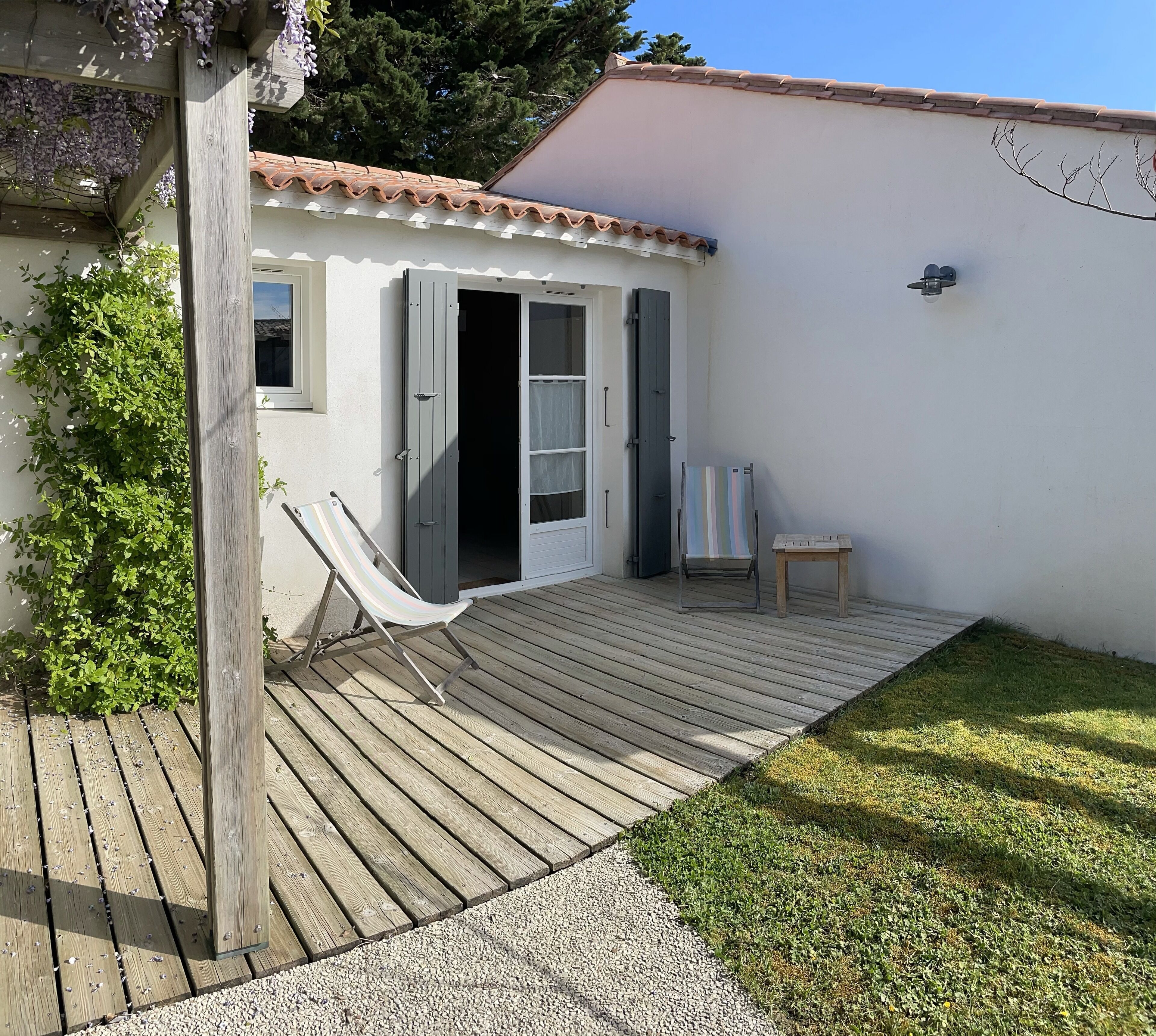 Una terraza de madera con una silla y una mesita, un edificio blanco con techo de tejas, y un arbusto verde.