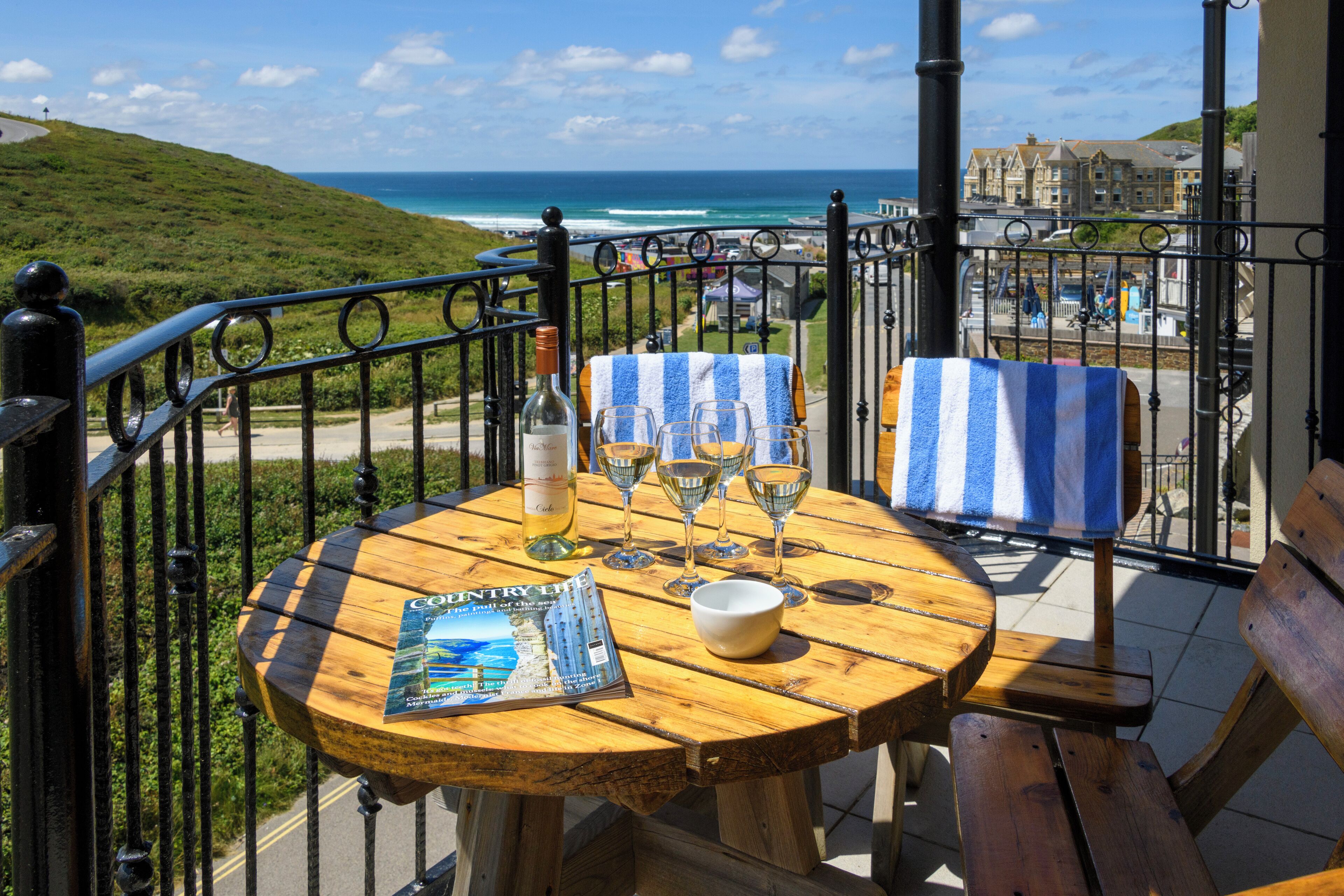 A wooden table with a magazine, wine glasses, and a bottle on a balcony overlooking the ocean.