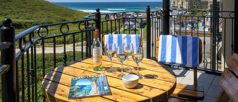 A wooden table with a magazine, wine glasses, and a bottle on a balcony overlooking the ocean.
