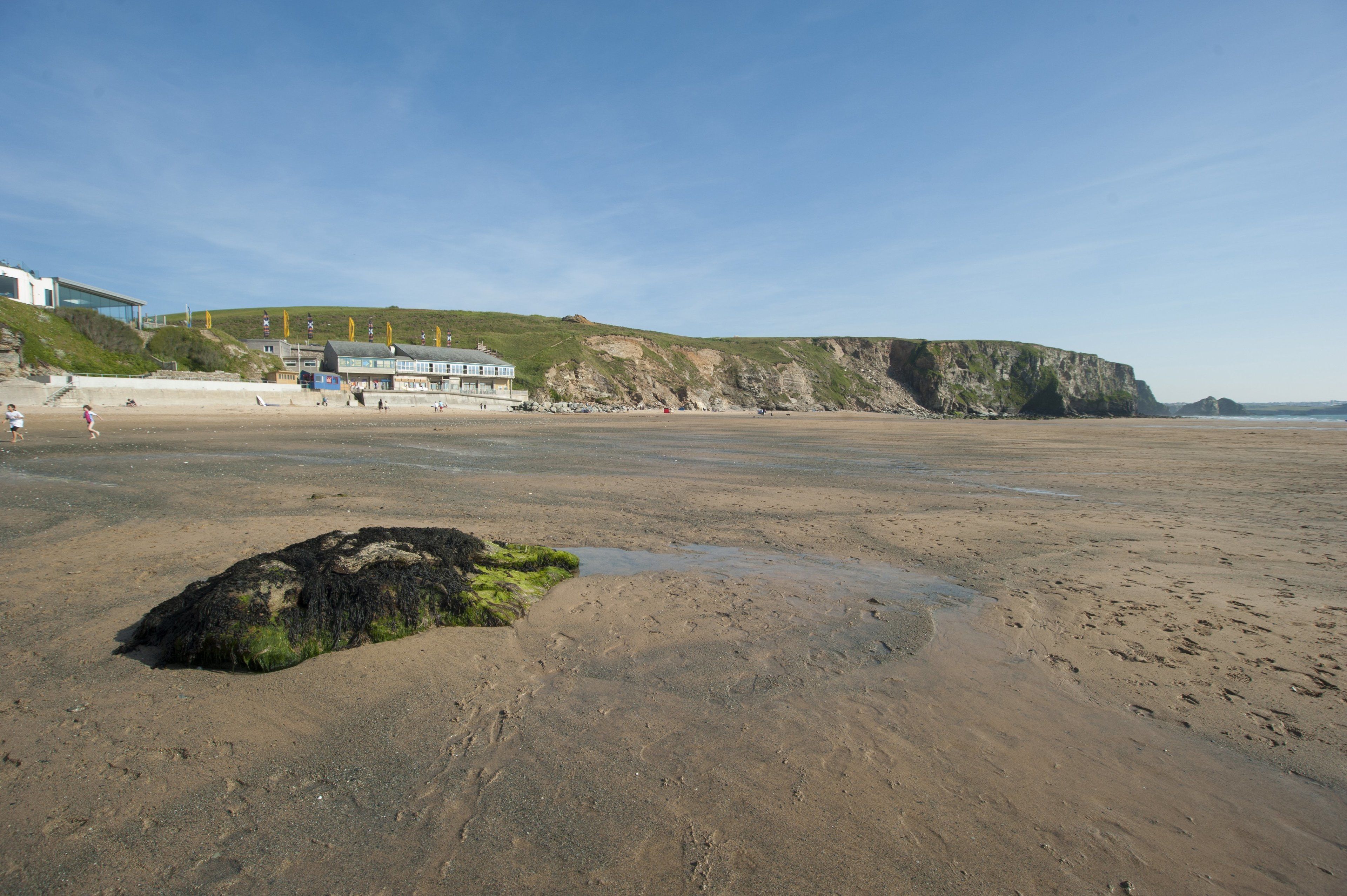 A beach with a building in the background, a cliff, and a large rock with green algae.