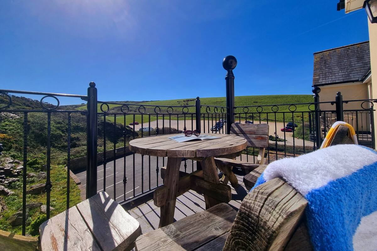 Un balcón con mesa y sillas de madera, barandilla de metal negro y vistas a un paisaje verde con colinas.