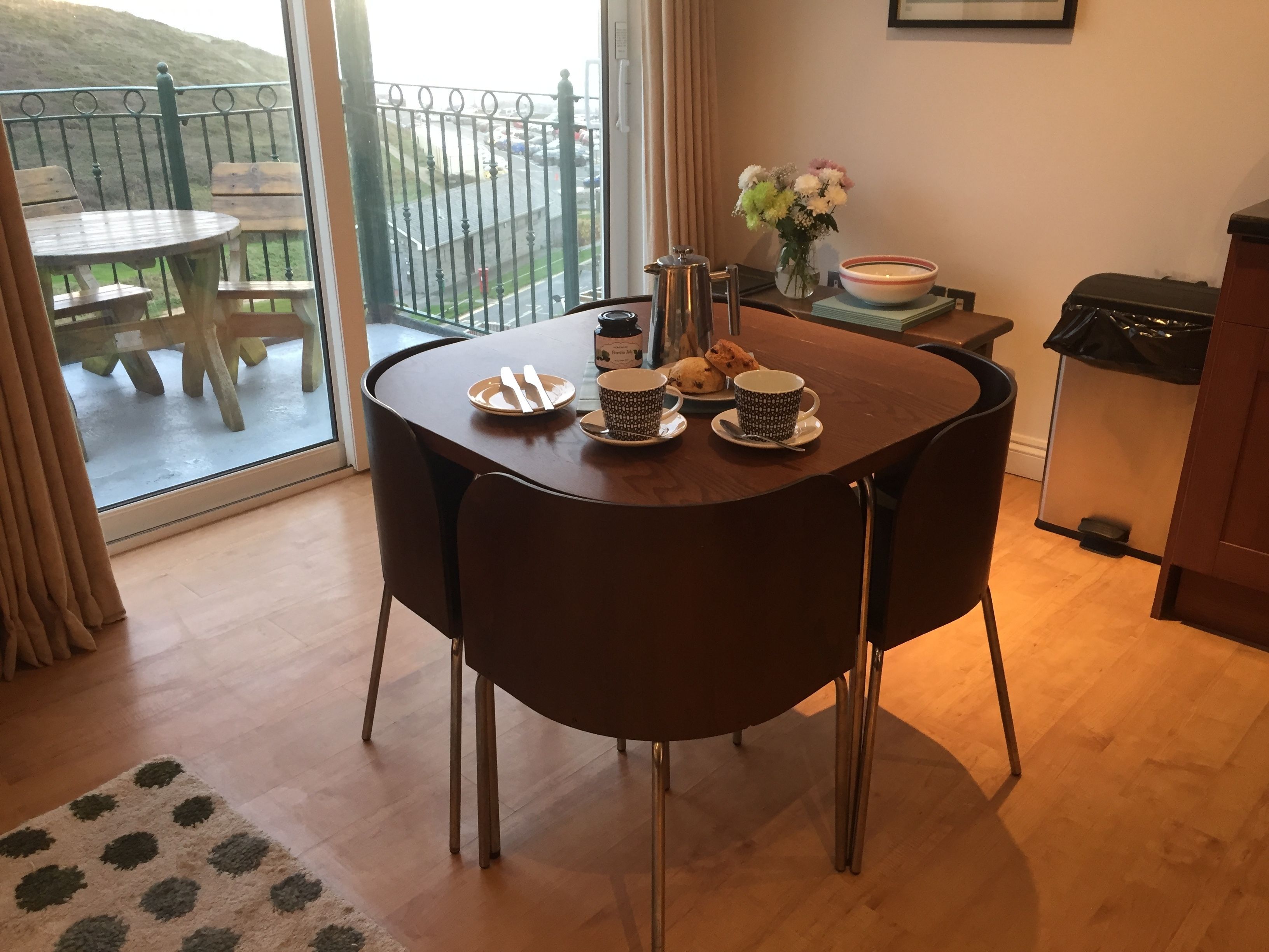 A dining area with a table set for two, a coffee pot, a bowl of cookies, and a vase of flowers.