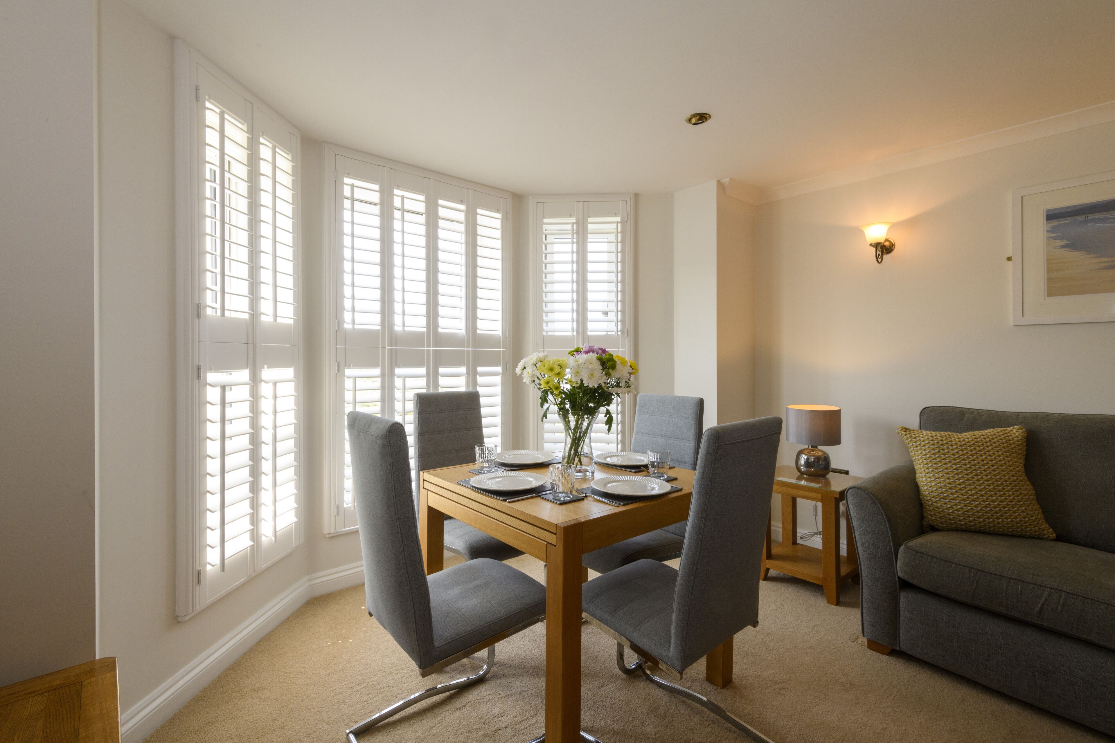 A dining area with a wooden table, four grey chairs, a vase with flowers, and a grey sofa in the background.