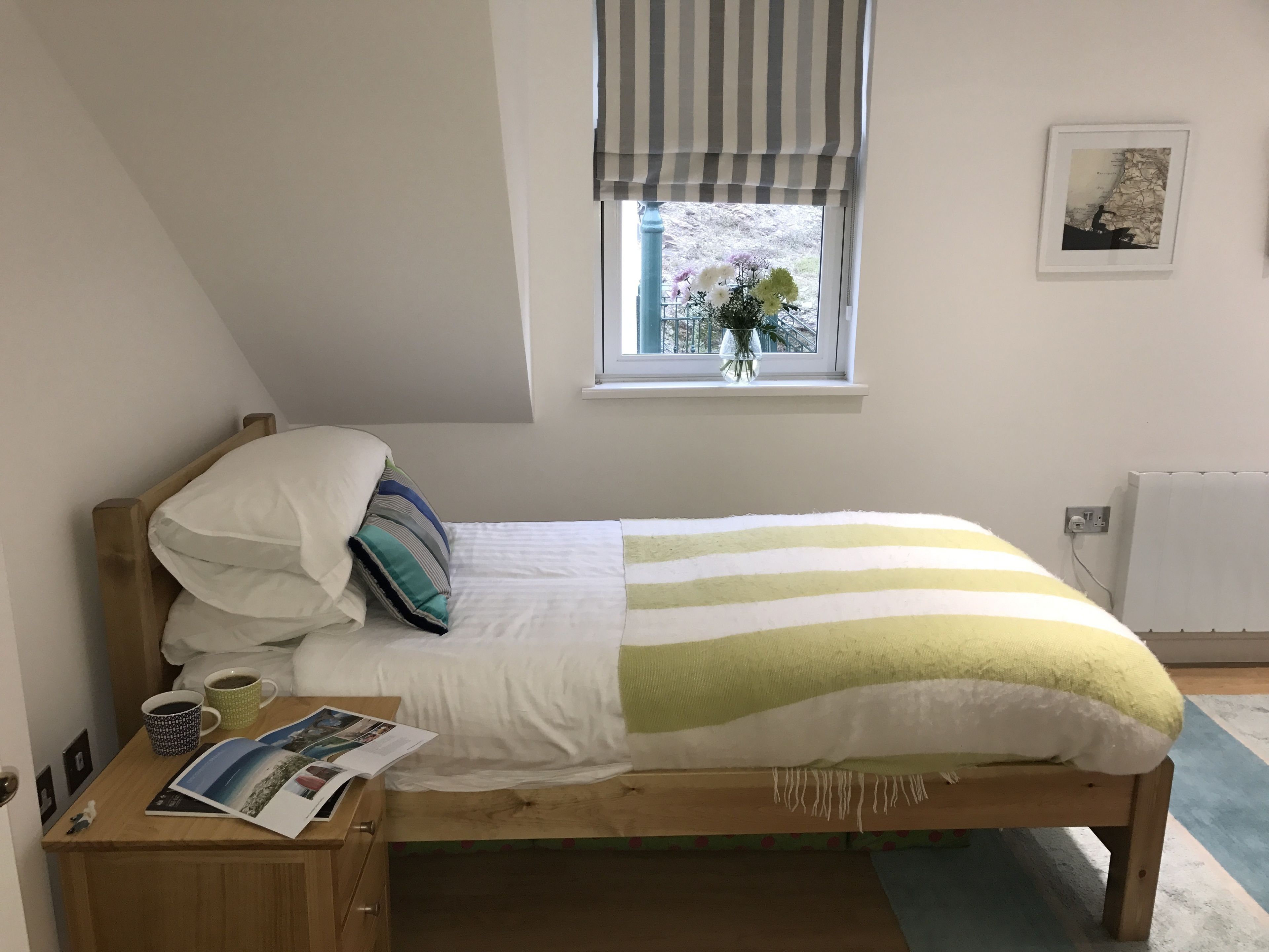 Een kleine, verzorgde slaapkamer met een enkel bed, een nachtkastje met een kop en tijdschrift, een raam met uitzicht op groen, en een radiator.