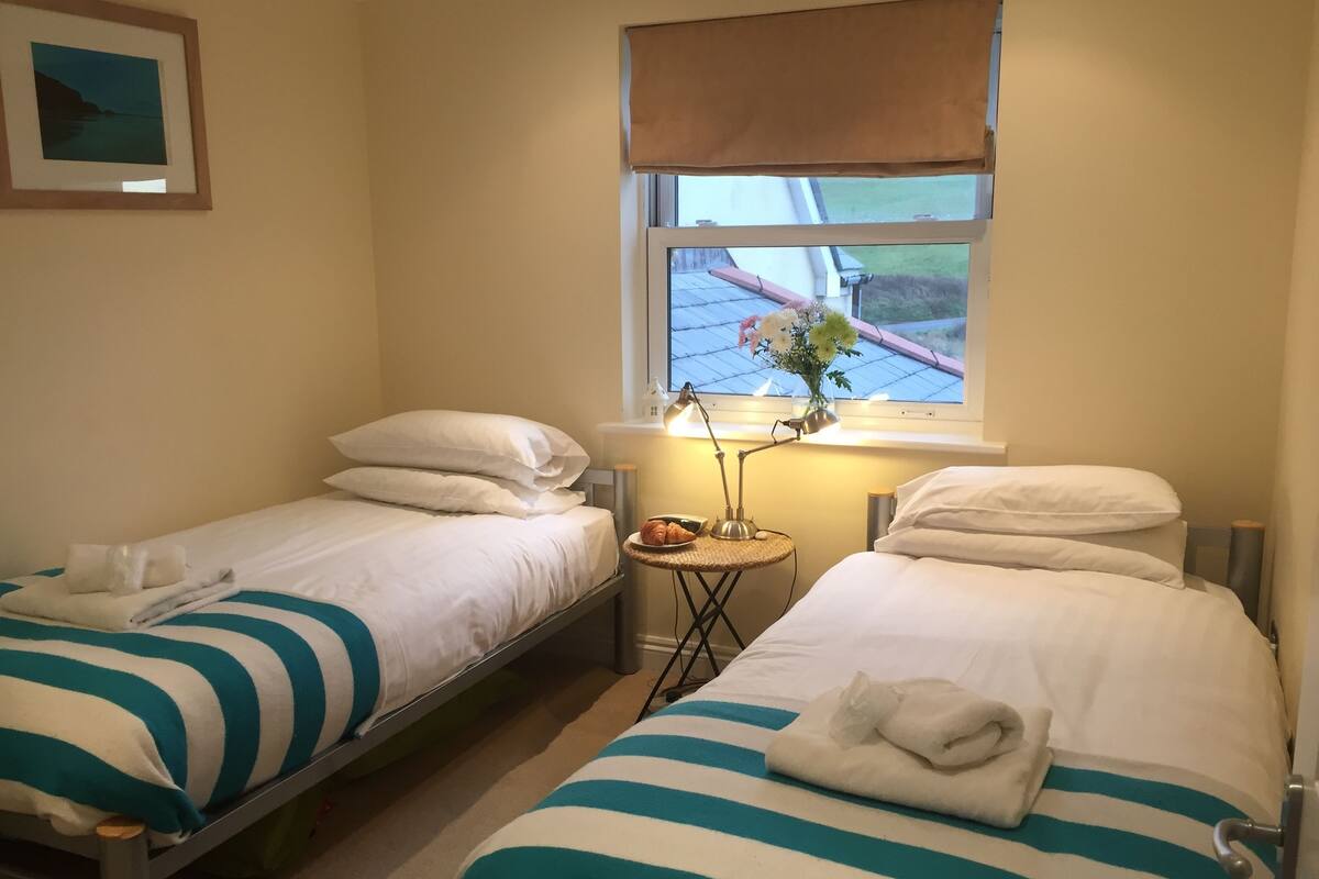 Habitación de hotel con dos camas individuales, cada una con una manta y almohadas a rayas. Hay una mesita con una lámpara y un florero con flores. Se ve una ventana con vistas a un edificio y una valla.