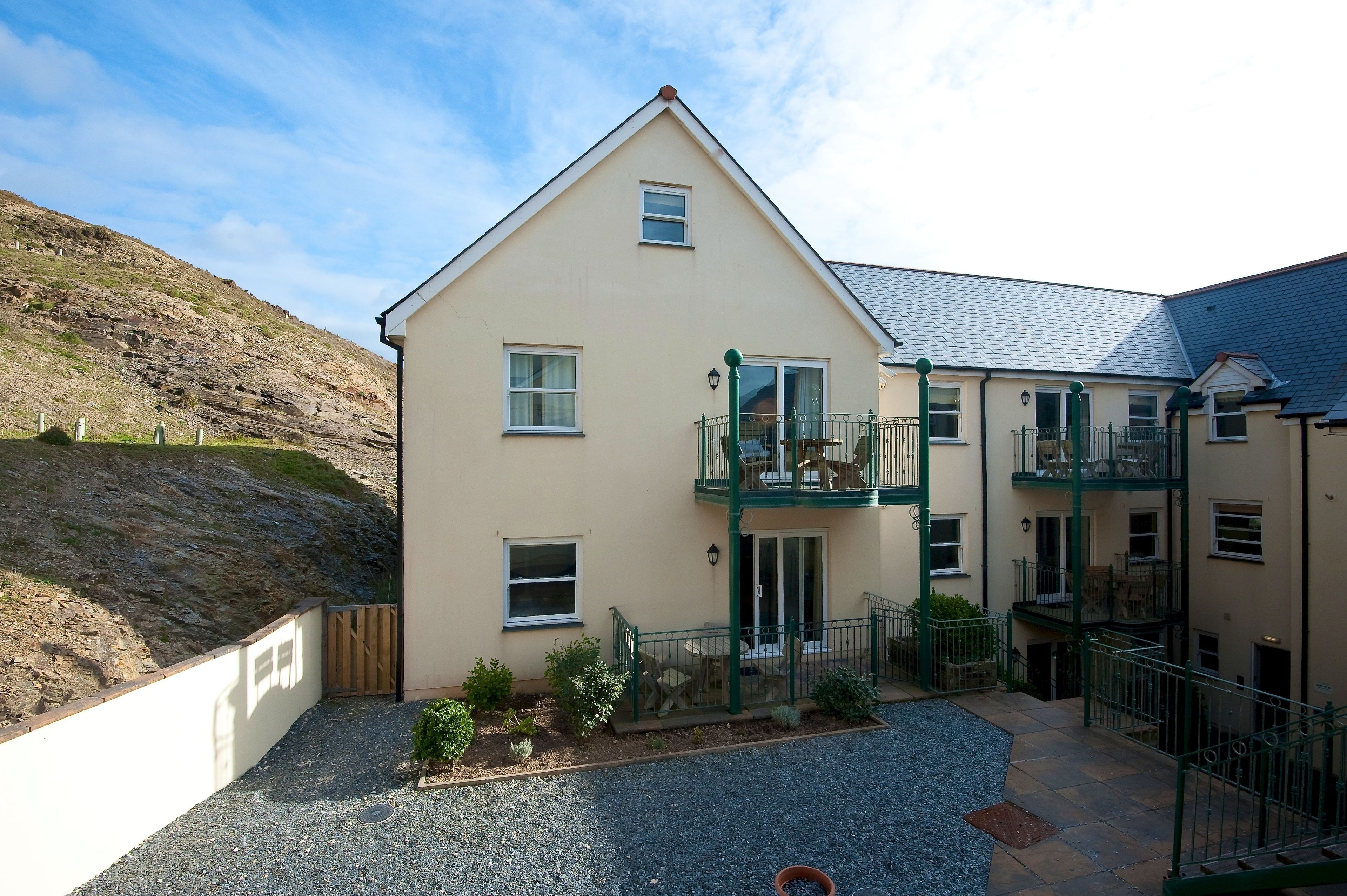 A two-story building with a balcony, a gravel driveway, and a hillside in the background.