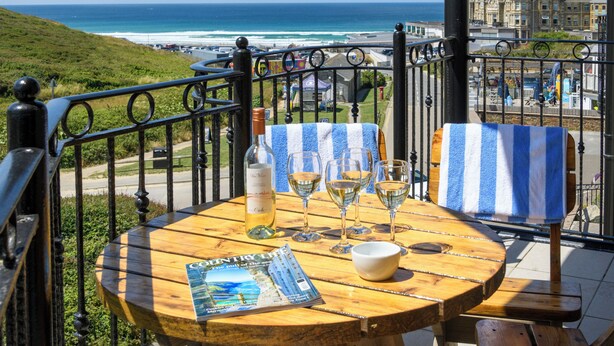 A wooden table with a magazine, a bottle, and wine glasses on a balcony overlooking a beach and buildings. - Beachcombers Apartments (Newquay)