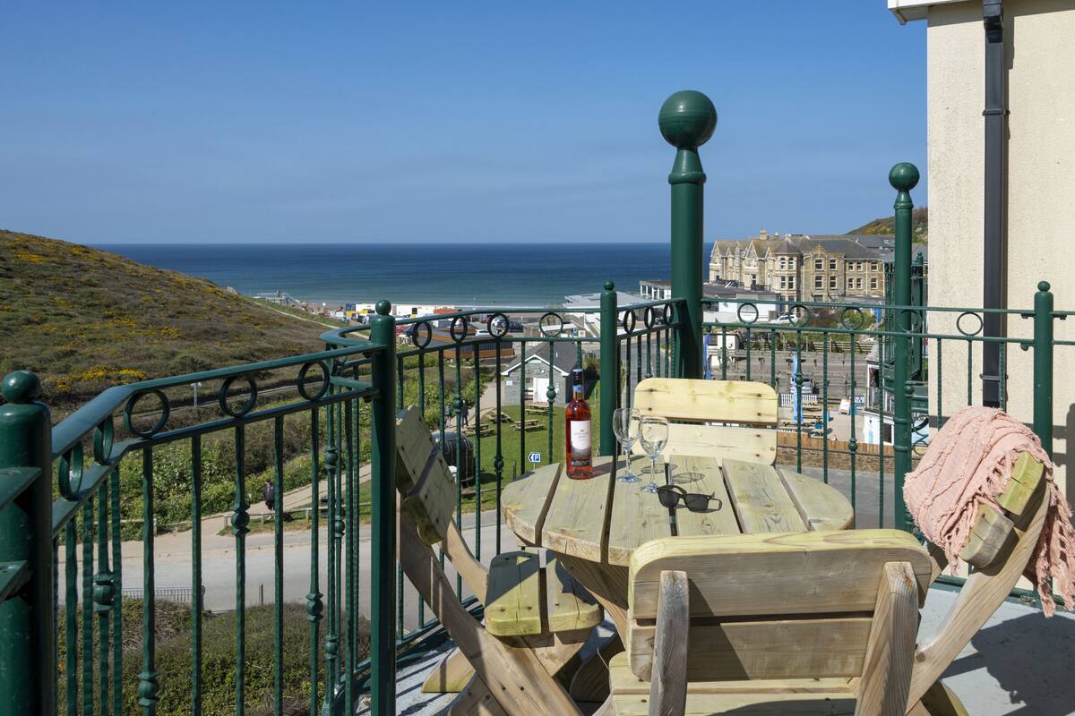 Una mesa de madera con una botella y vasos, una barandilla verde, y una vista al mar y edificios al fondo.