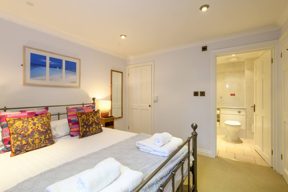 Un dormitorio ordenado con cama, almohadas, una mesita de noche con lámpara, una puerta que da a un baño y un cuadro en la pared.