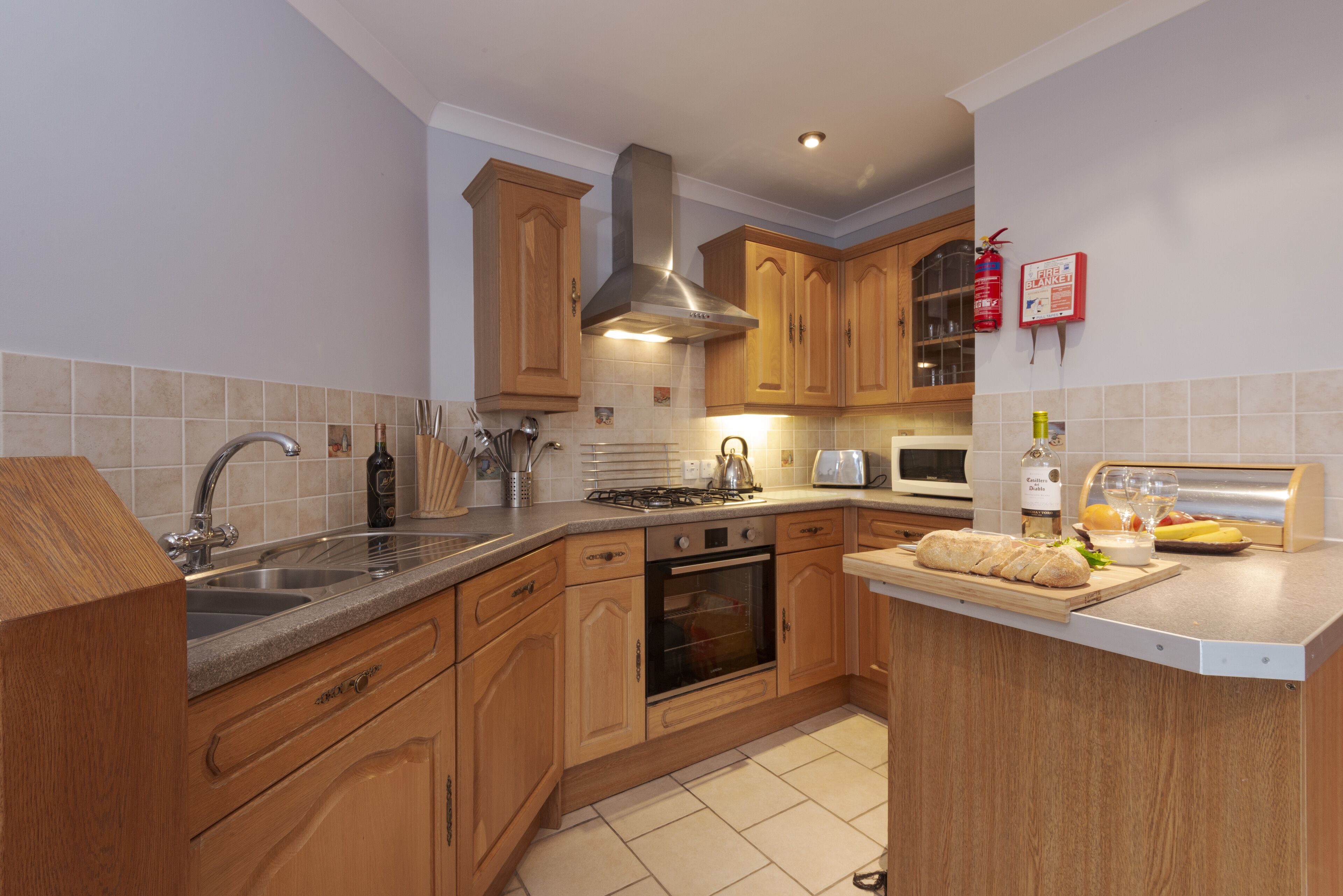 A kitchen with wooden cabinets, a stainless steel range hood, a built-in oven, and a countertop with a cutting board and bread.