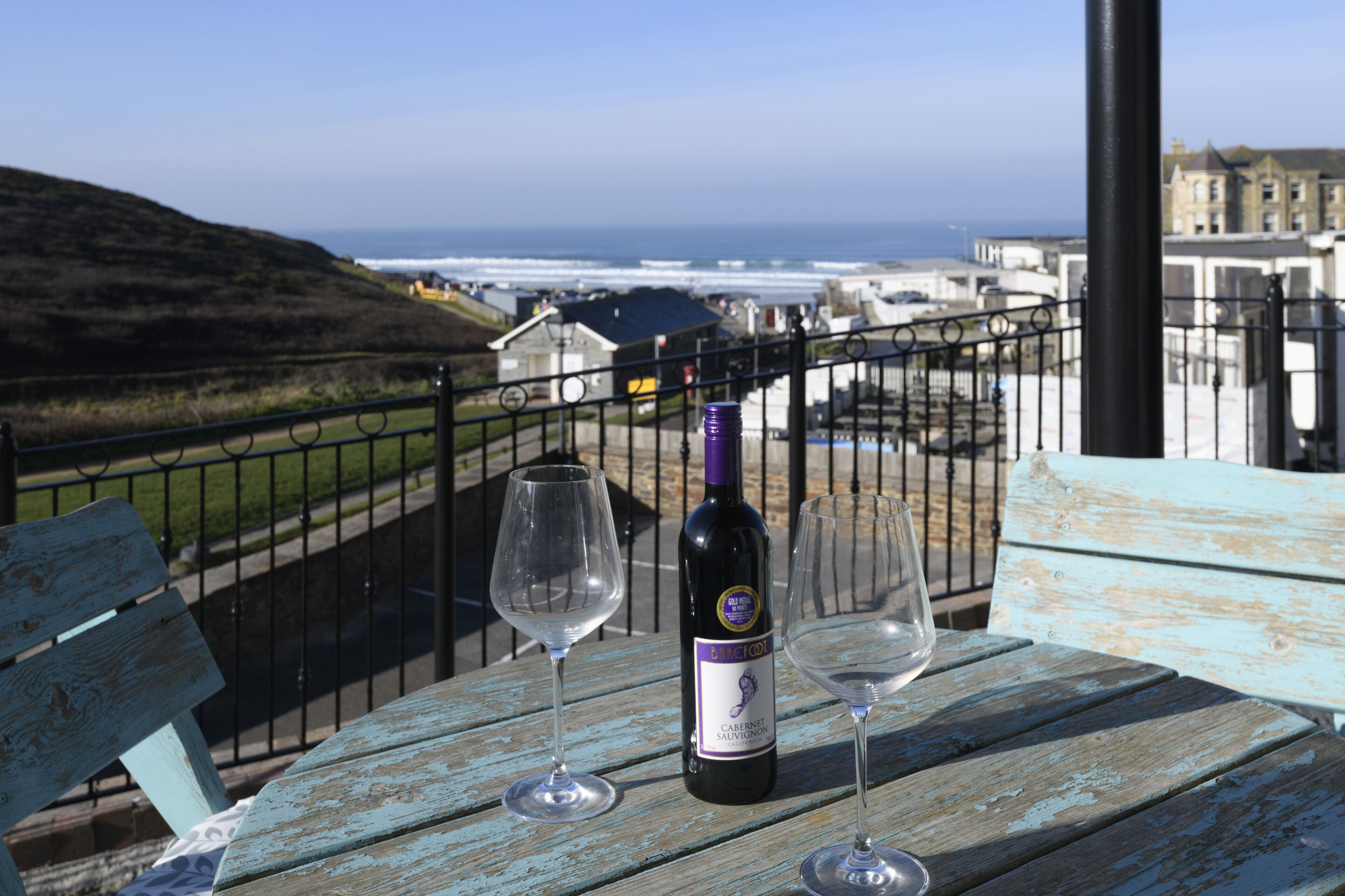 A table with a bottle of wine and two glasses on a balcony overlooking a coastal view.