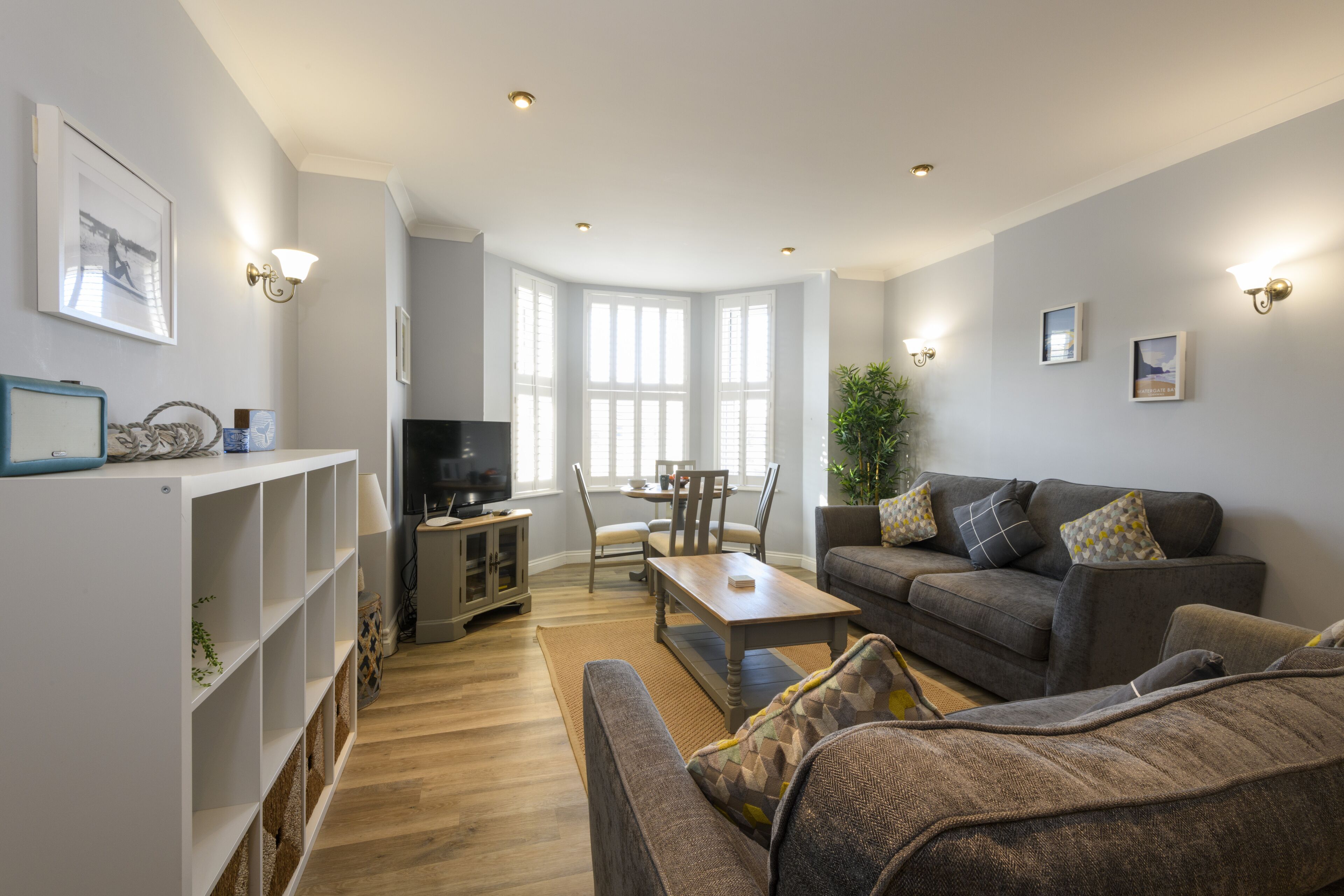 A living room with a sofa, a coffee table, and a TV stand. There is a dining table with chairs and a plant.