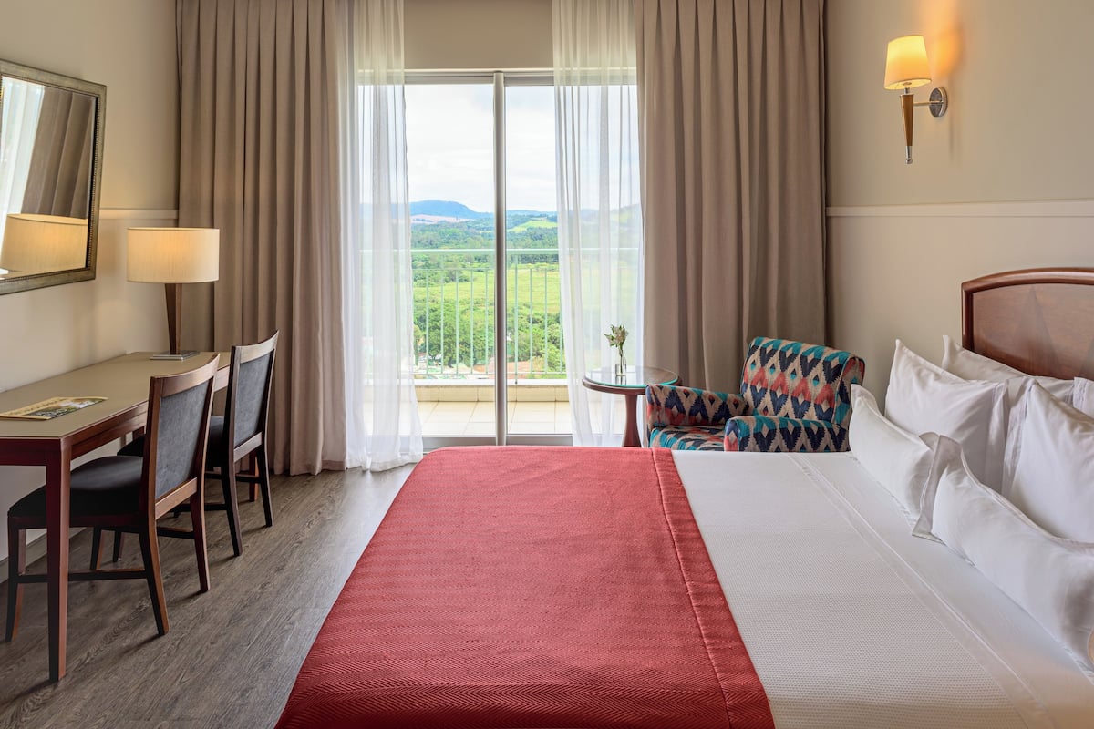 A hotel room with a large bed, a desk with chairs, a small table, and a view of a green landscape through the window.