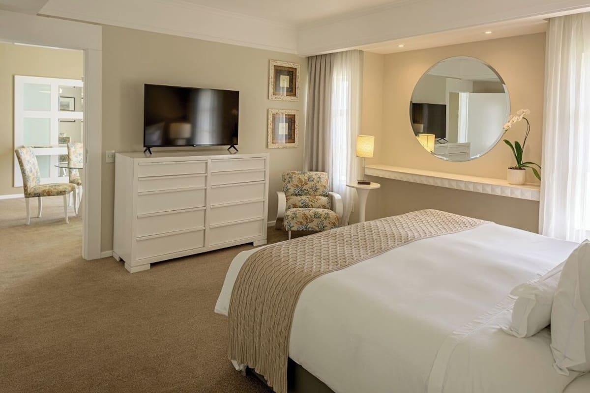 A hotel room with a large bed, a TV on a dresser, a chair, a round mirror, and a small table with a lamp.