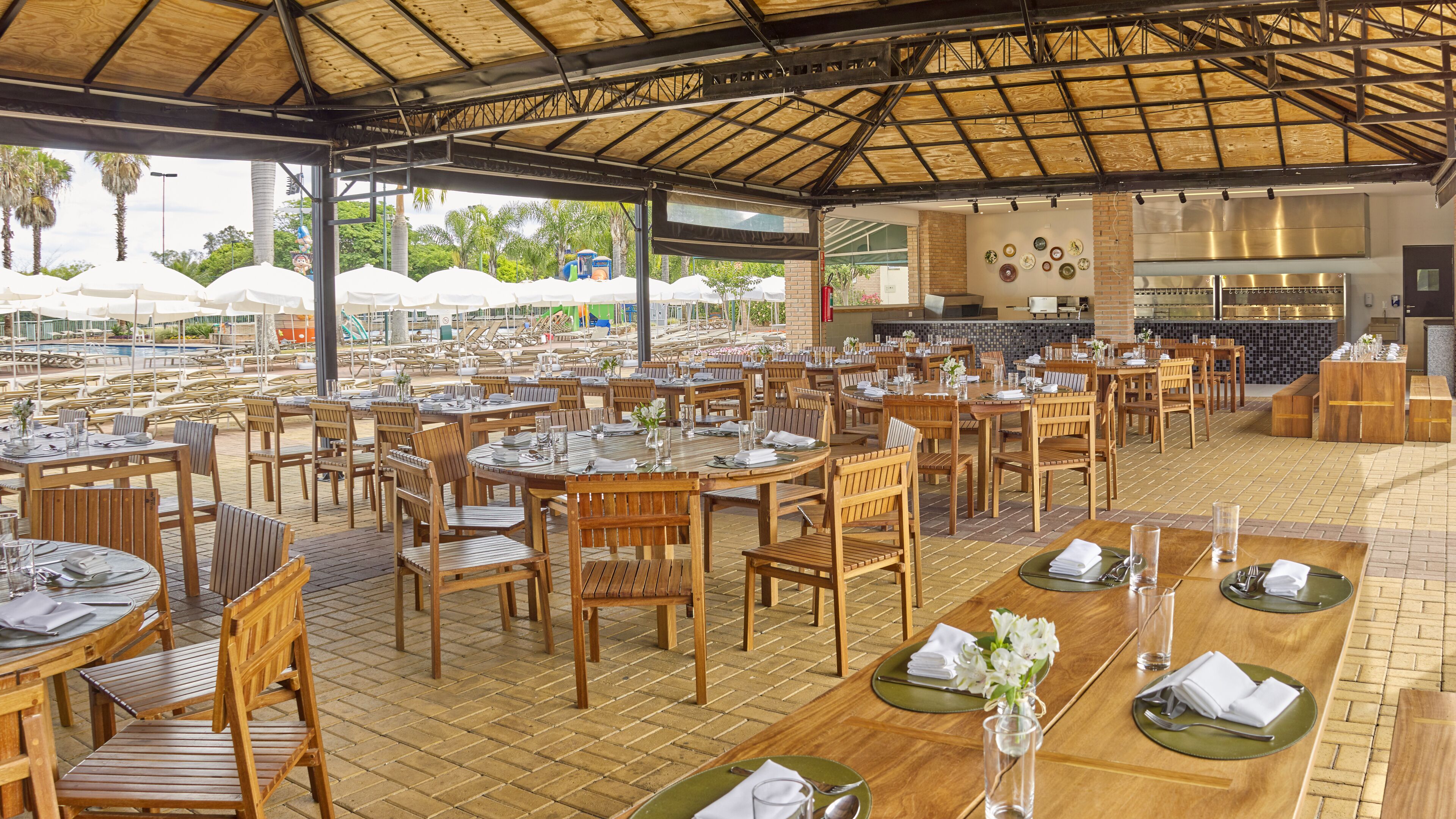 An outdoor dining area with multiple tables and chairs, a covered section with a wooden ceiling, and a view of a pool and palm trees in the background.