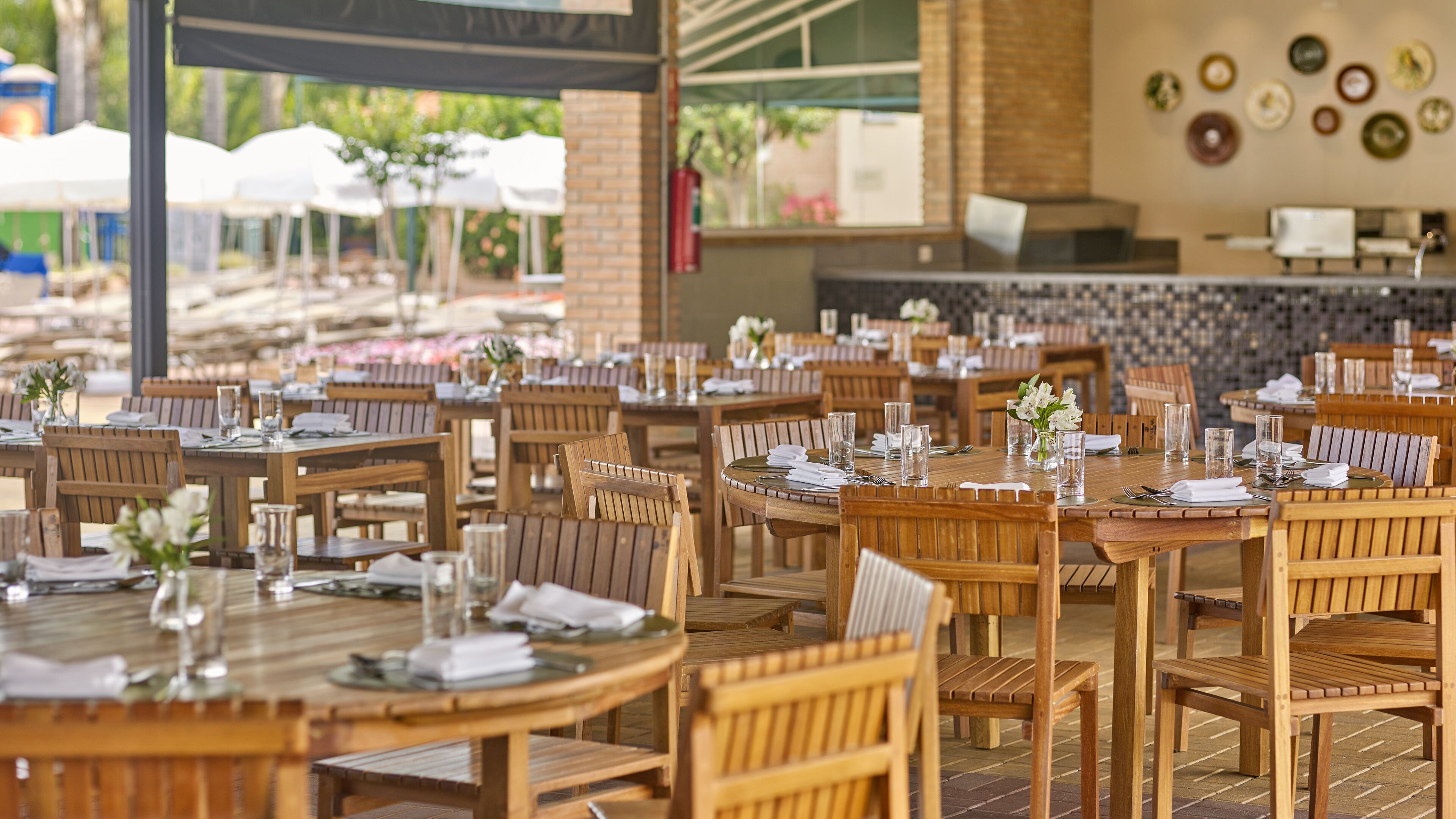 A restaurant with wooden tables and chairs, arranged in a spacious outdoor setting with brick walls and a tiled floor.
