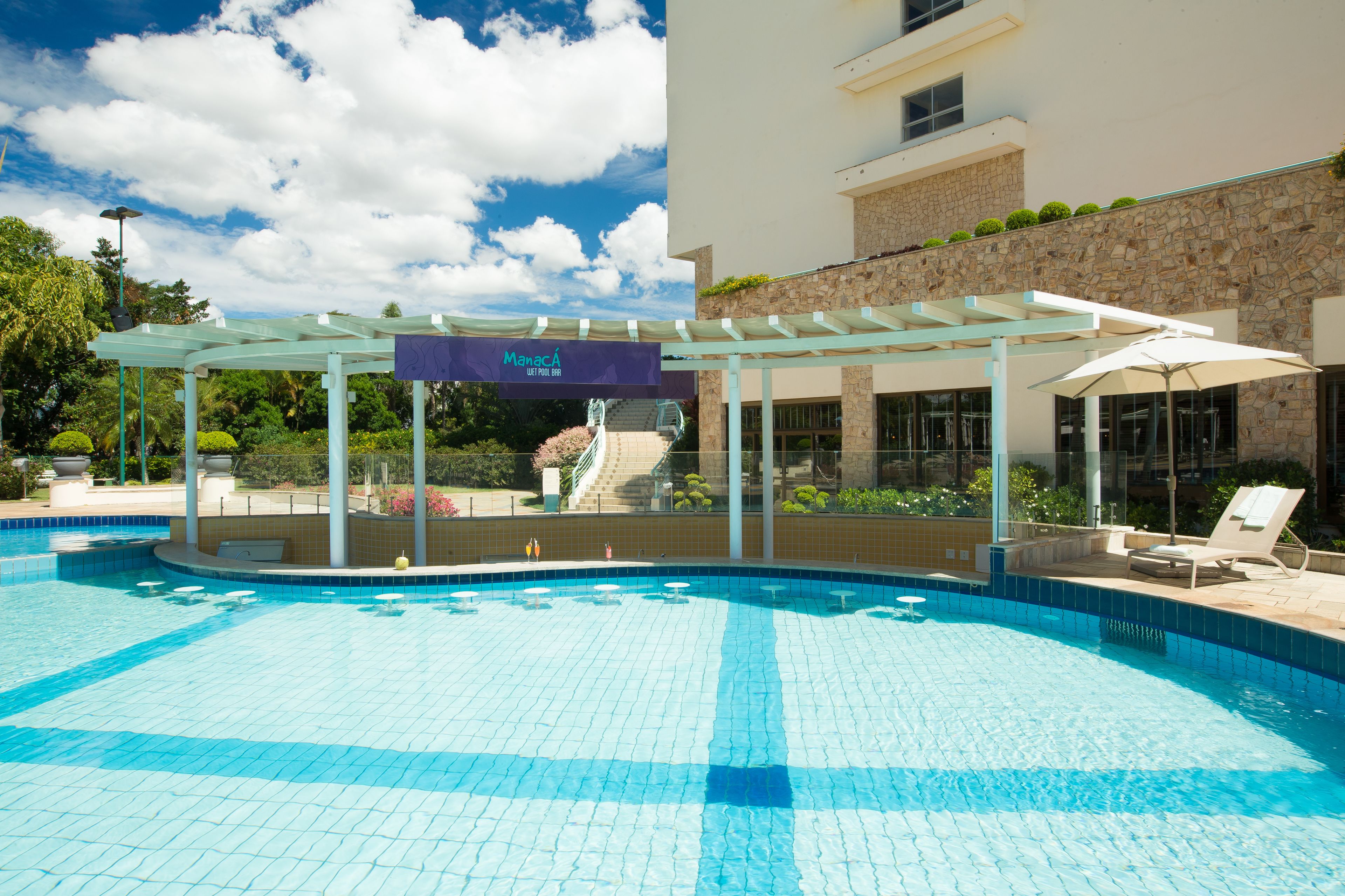 Una piscina con zona cubierta, un cartel que dice "Manac" y un edificio de fachada de piedra y vegetación.