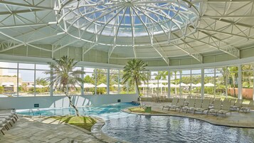 Piscina interna com teto de vidro, espreguiçadeiras e palmeiras.