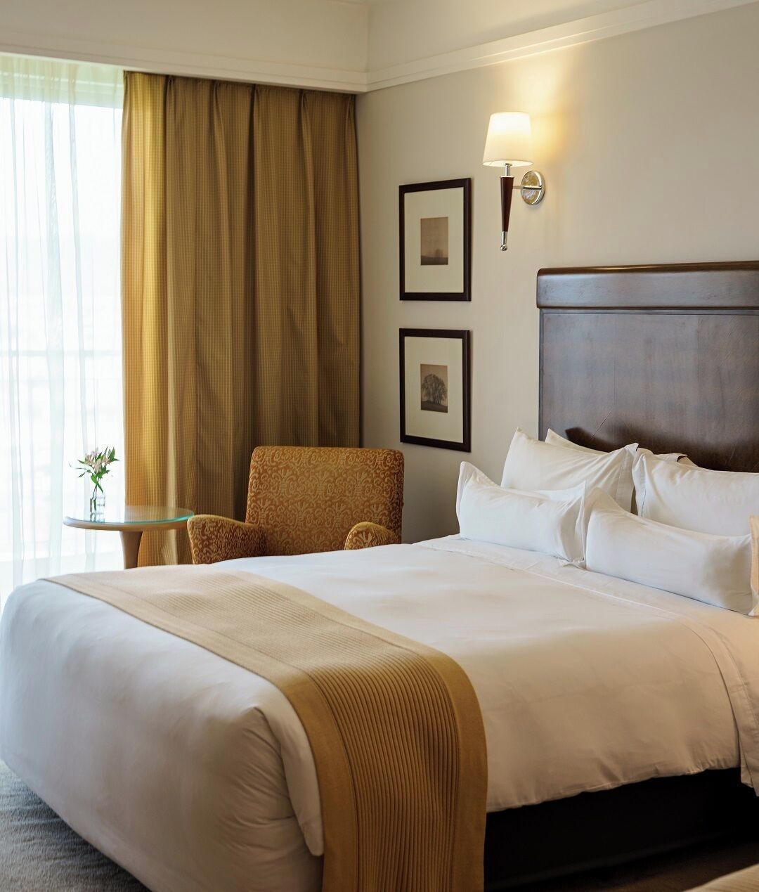 A hotel room with a large bed, a chair, a small table with a vase, and two framed pictures on the wall.