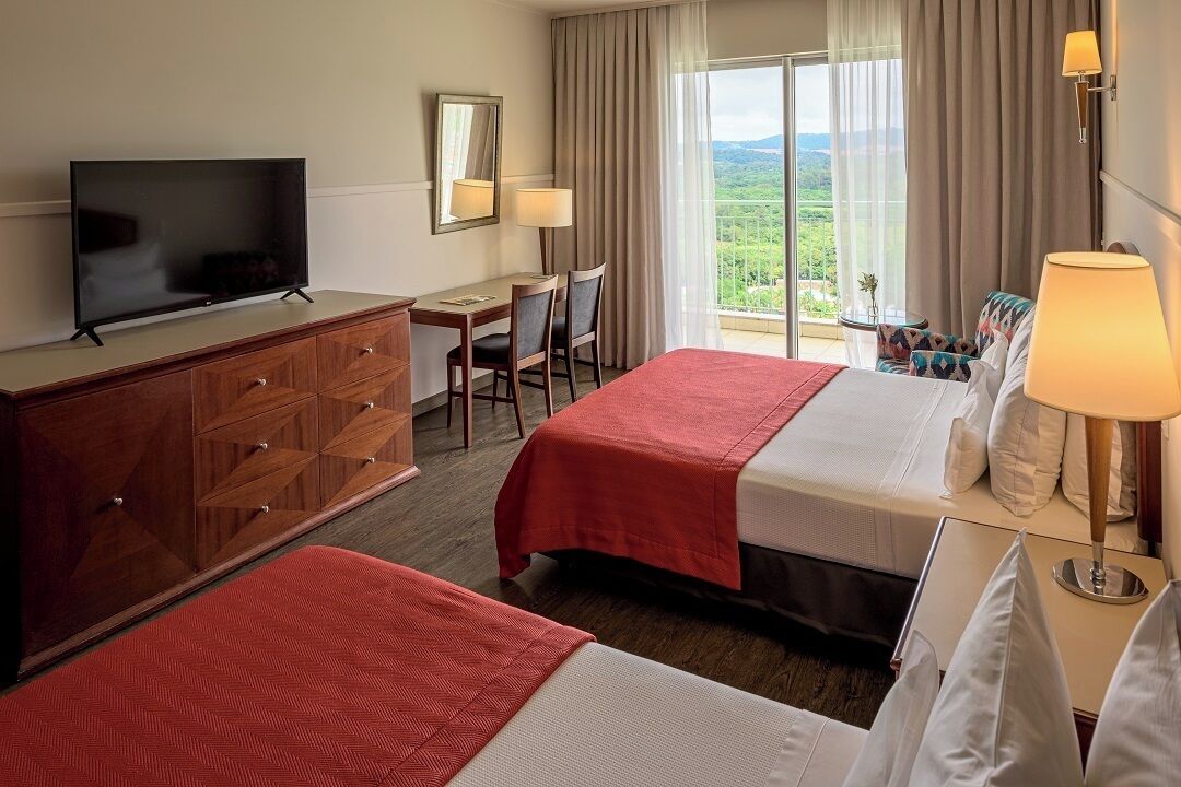 Habitación de hotel con dos camas, televisión, escritorio con sillas, lámpara y vistas al exterior.