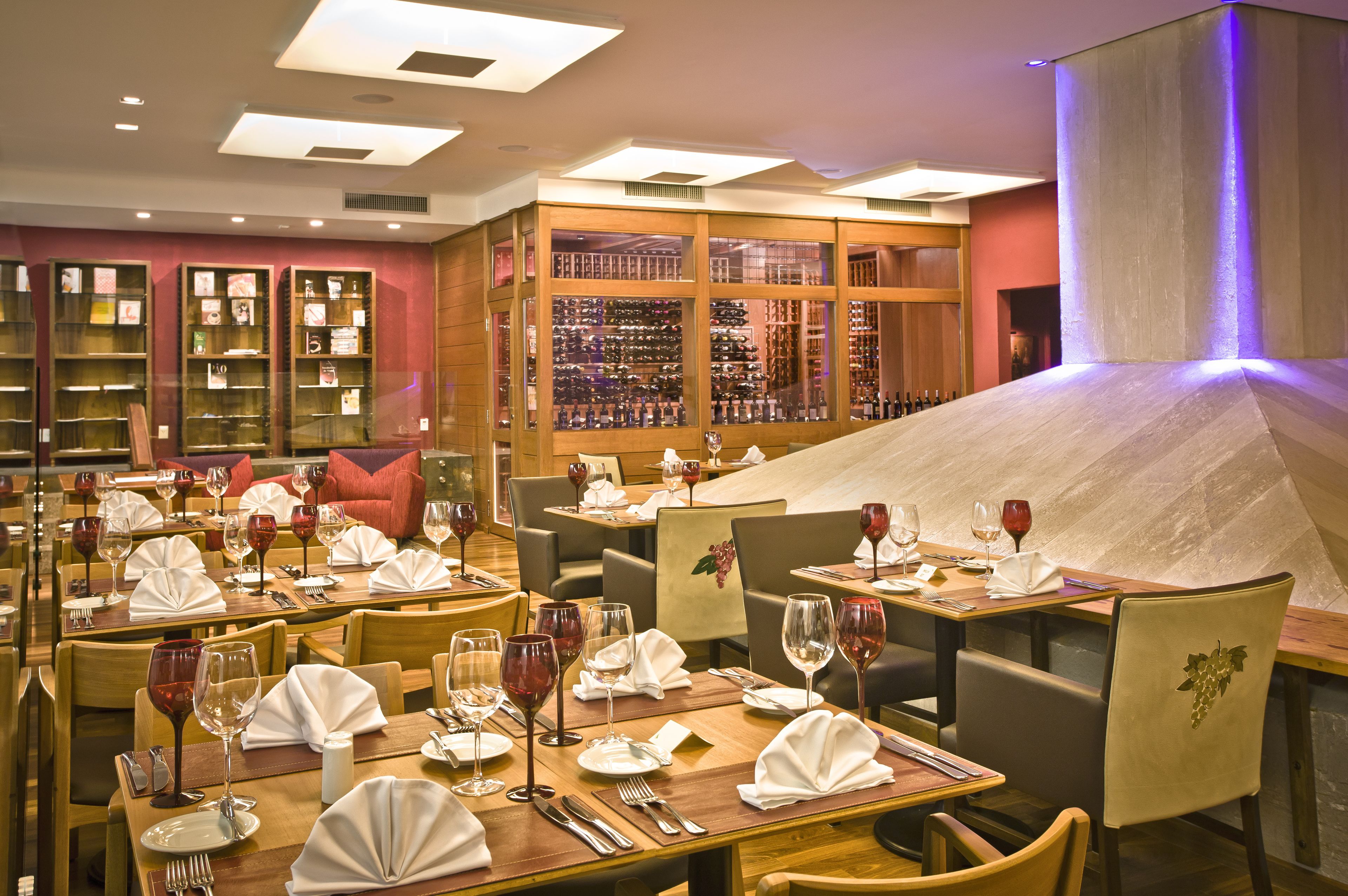 A restaurant with multiple dining tables set with wine glasses, napkins, and cutlery. There are bookshelves and a wine rack in the background.