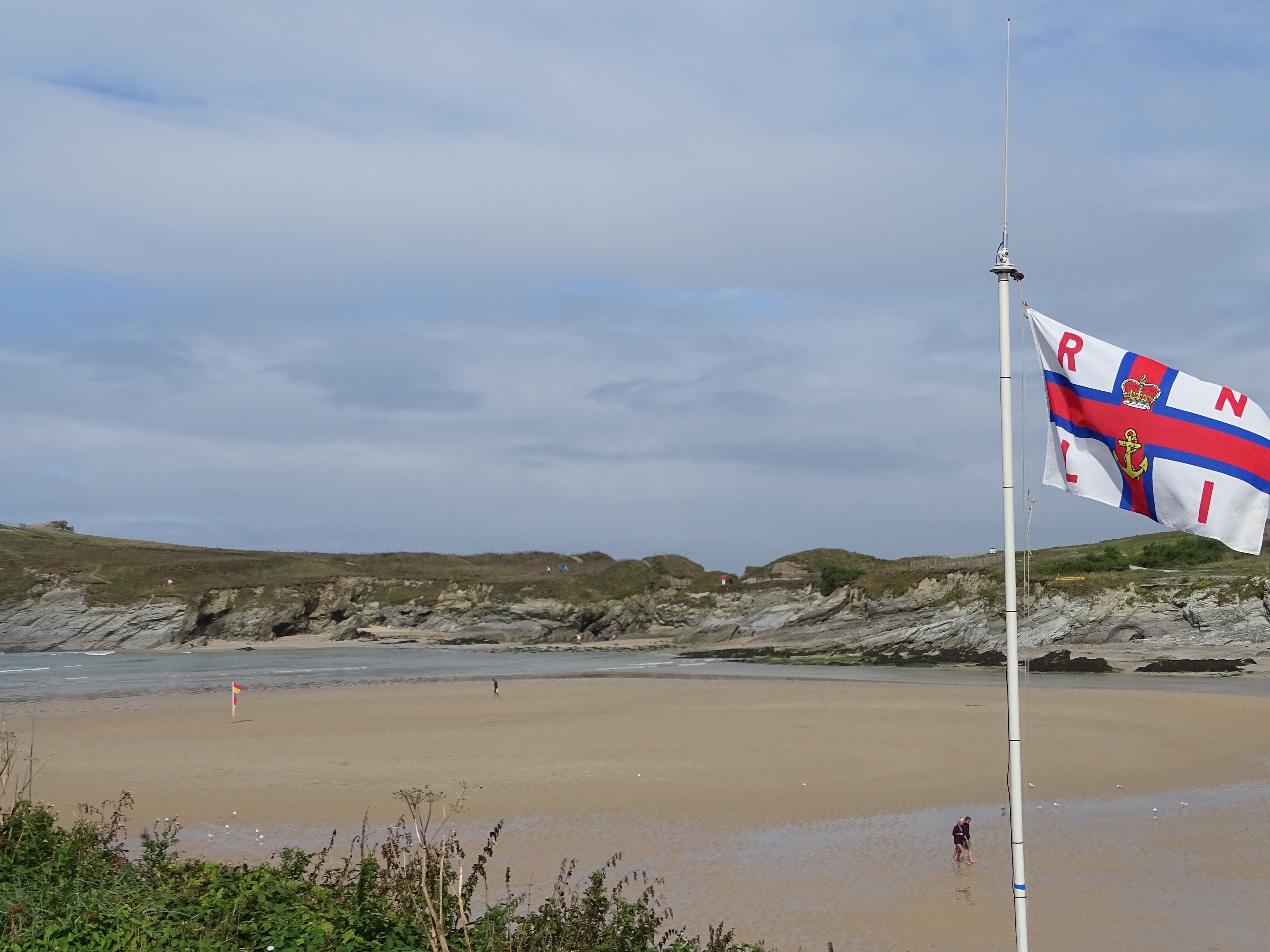 Una playa con un asta de bandera ondeando una enseña con el emblema de una corona y un ancla.