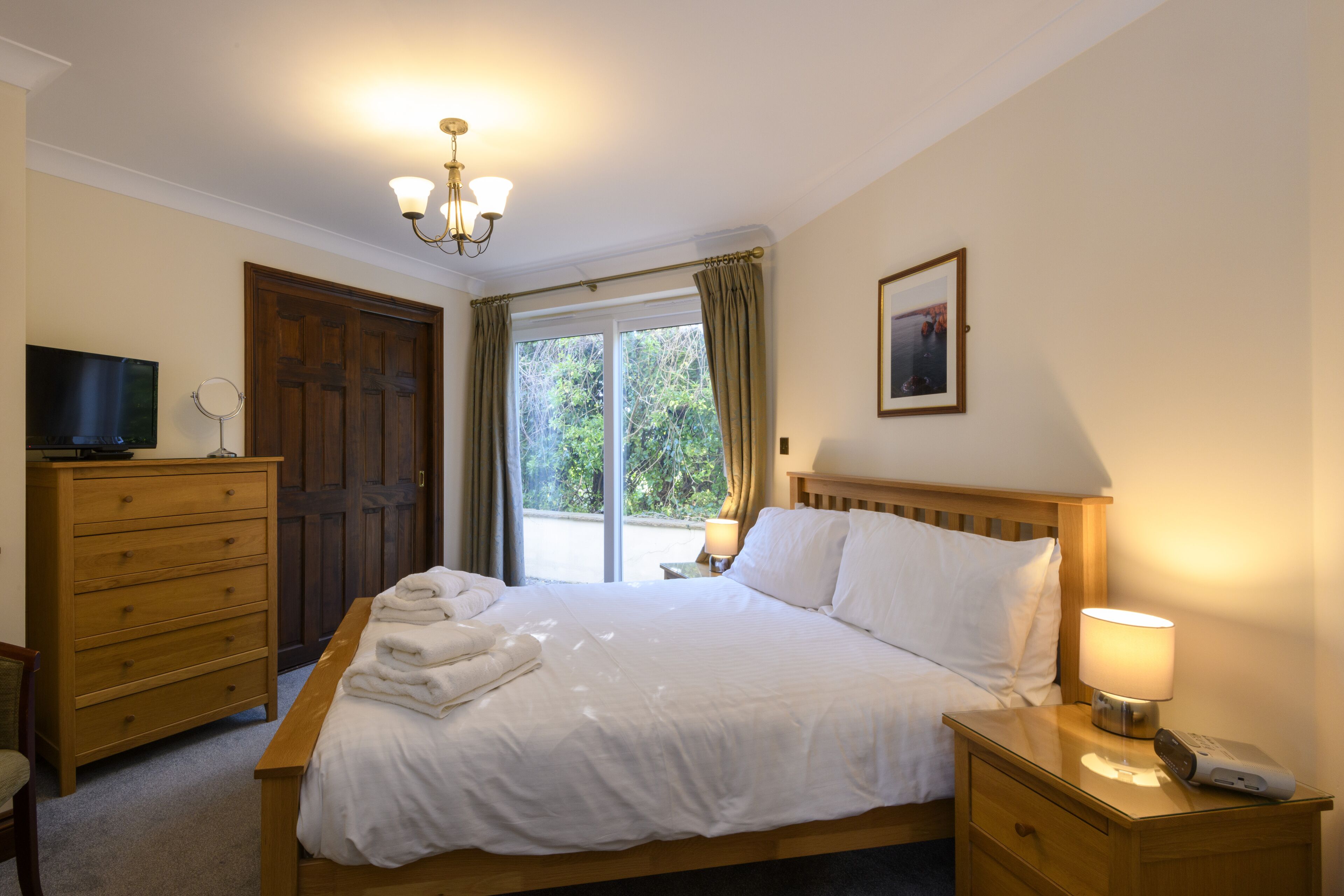 A neatly made bed with white linens, a wooden nightstand with a lamp, a dresser, and a television on the left.