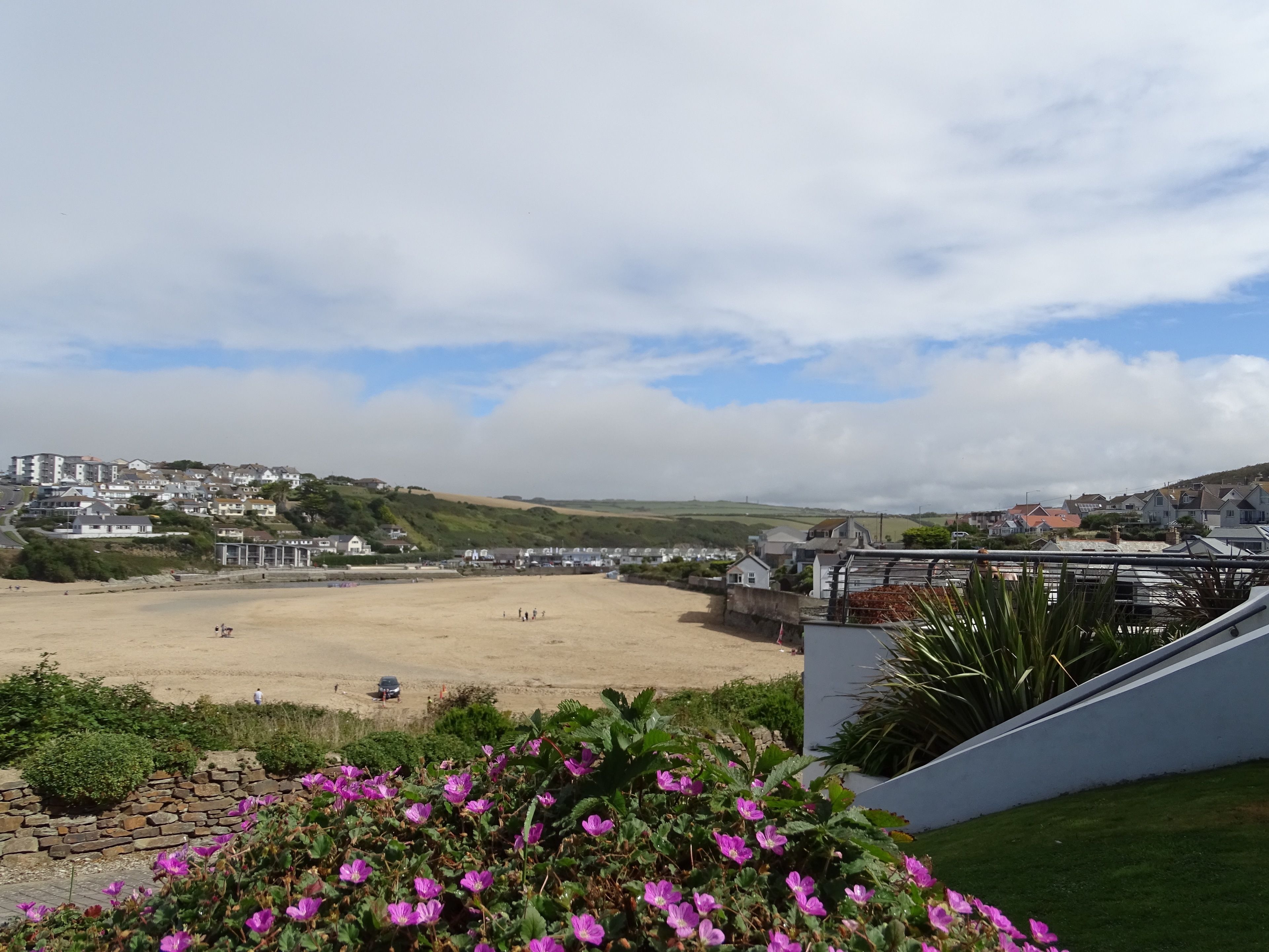 Vista costera con una playa arenosa, una zona cubierta de césped con flores moradas y un pueblo al fondo.