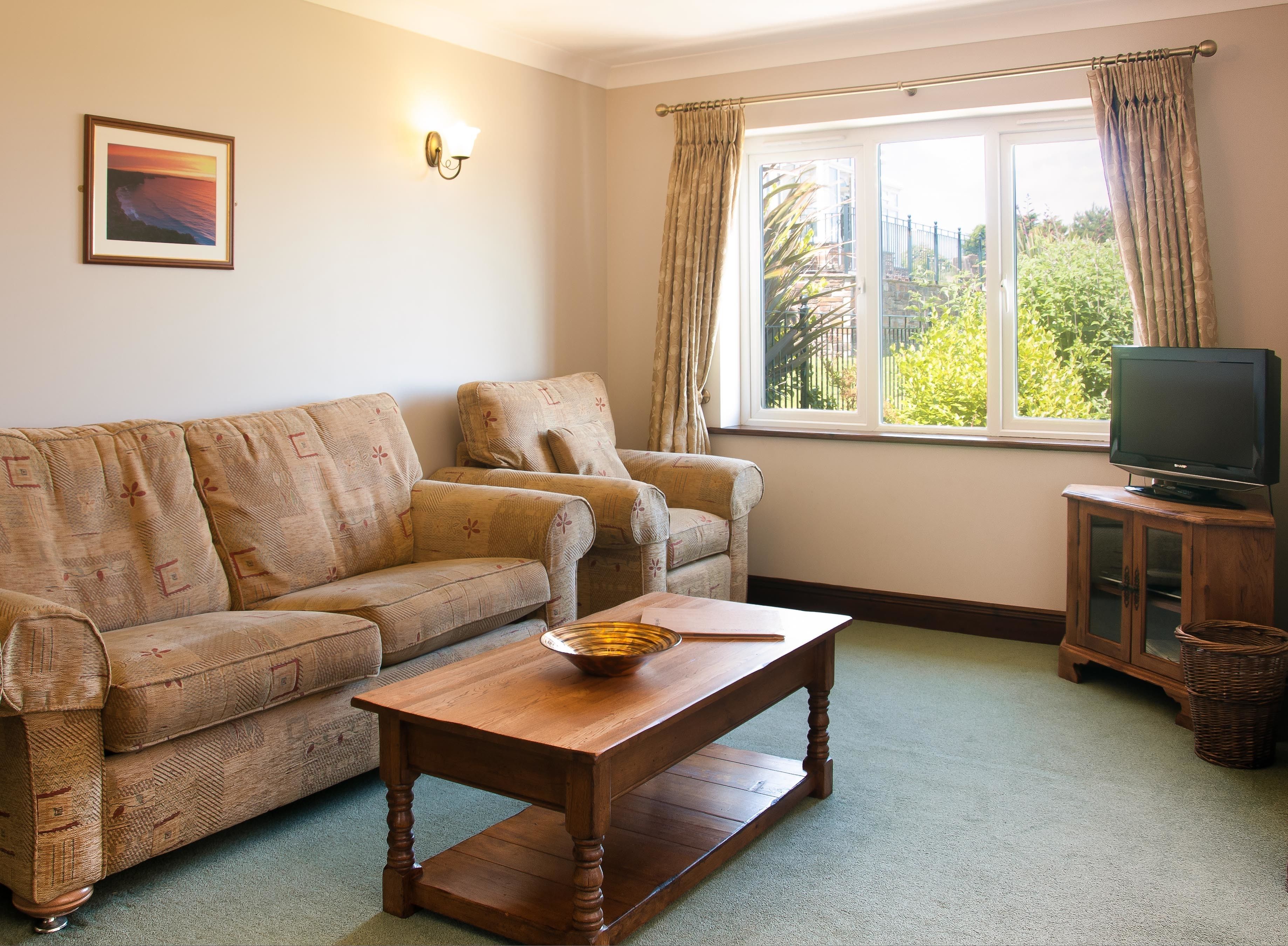 A living room with a sofa, armchair, wooden coffee table, television on a cabinet, and a window with curtains.