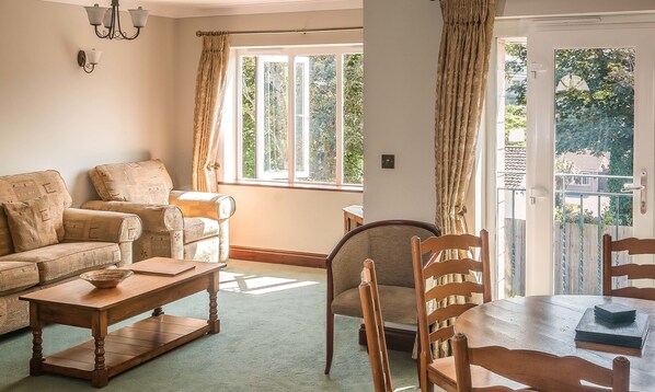 A living room with a sofa, a wooden coffee table, and a dining table with chairs. There are large windows with curtains, a chandelier, and a view of greenery outside. - Porth Veor Villas & Apartments (Newquay)