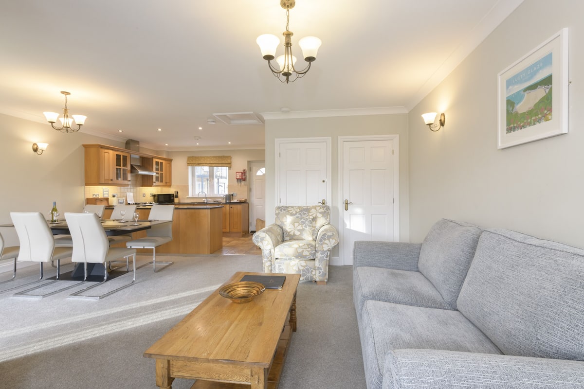 A spacious living area with a grey sofa, a wooden coffee table, and a patterned armchair. A kitchen with wooden cabinets and a dining area with white chairs is visible in the background.