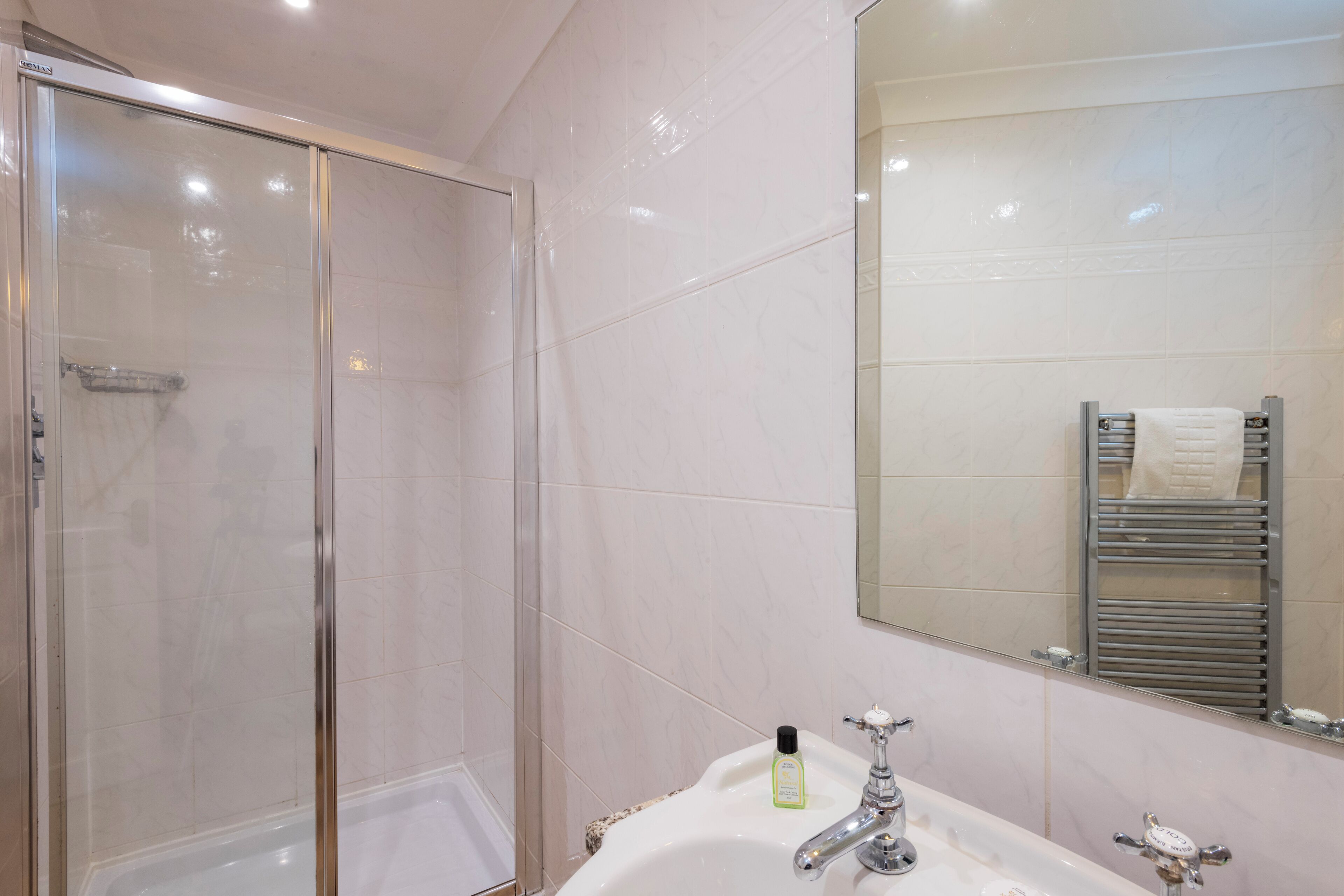 Un baño con ducha envuelta en vidrio, un lavamanos blanco con un espejo encima y un perchero con una toalla.