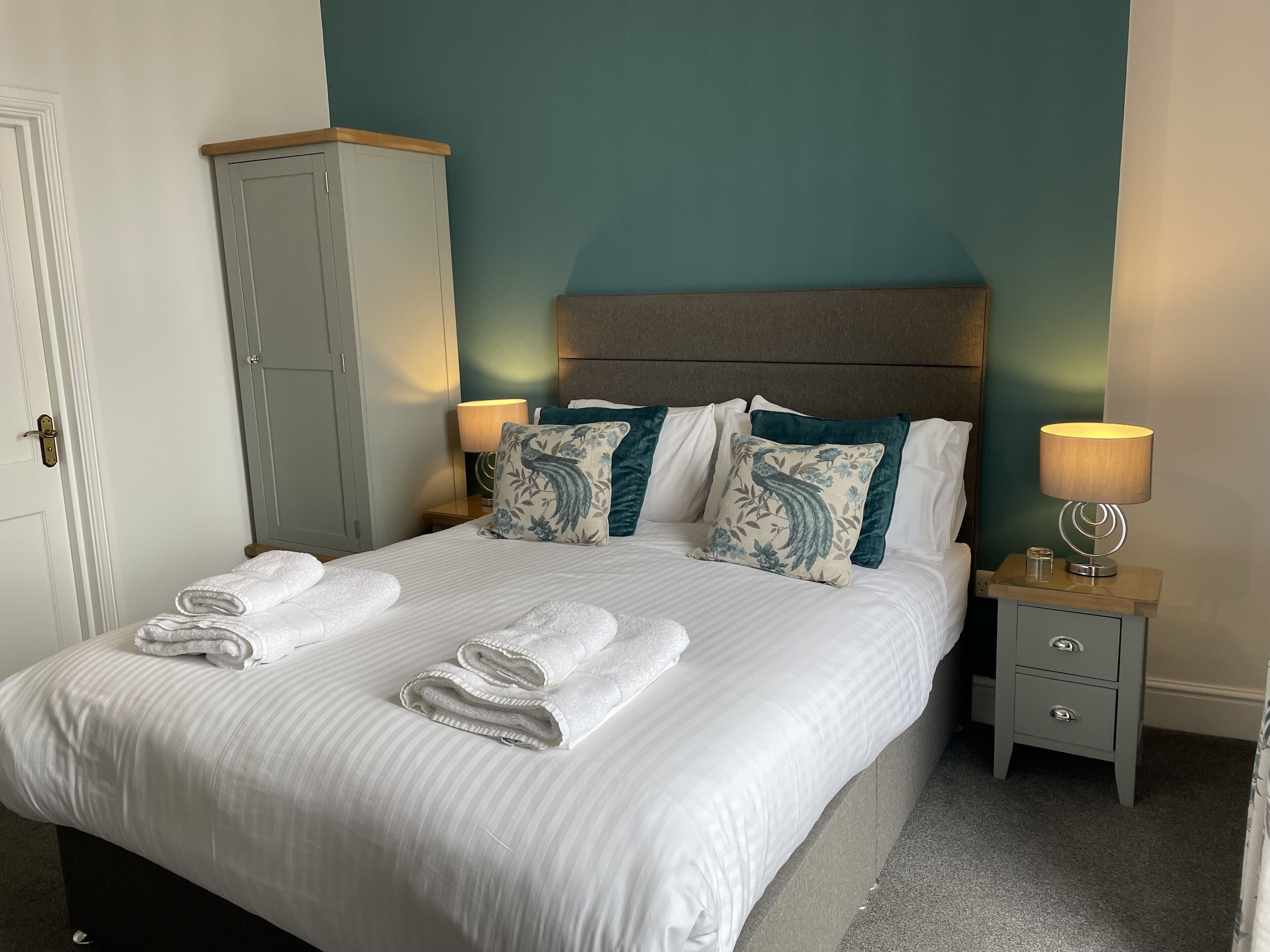 A neatly made bed with white linens and rolled towels, a wooden headboard, teal walls, and two bedside tables with lamps.