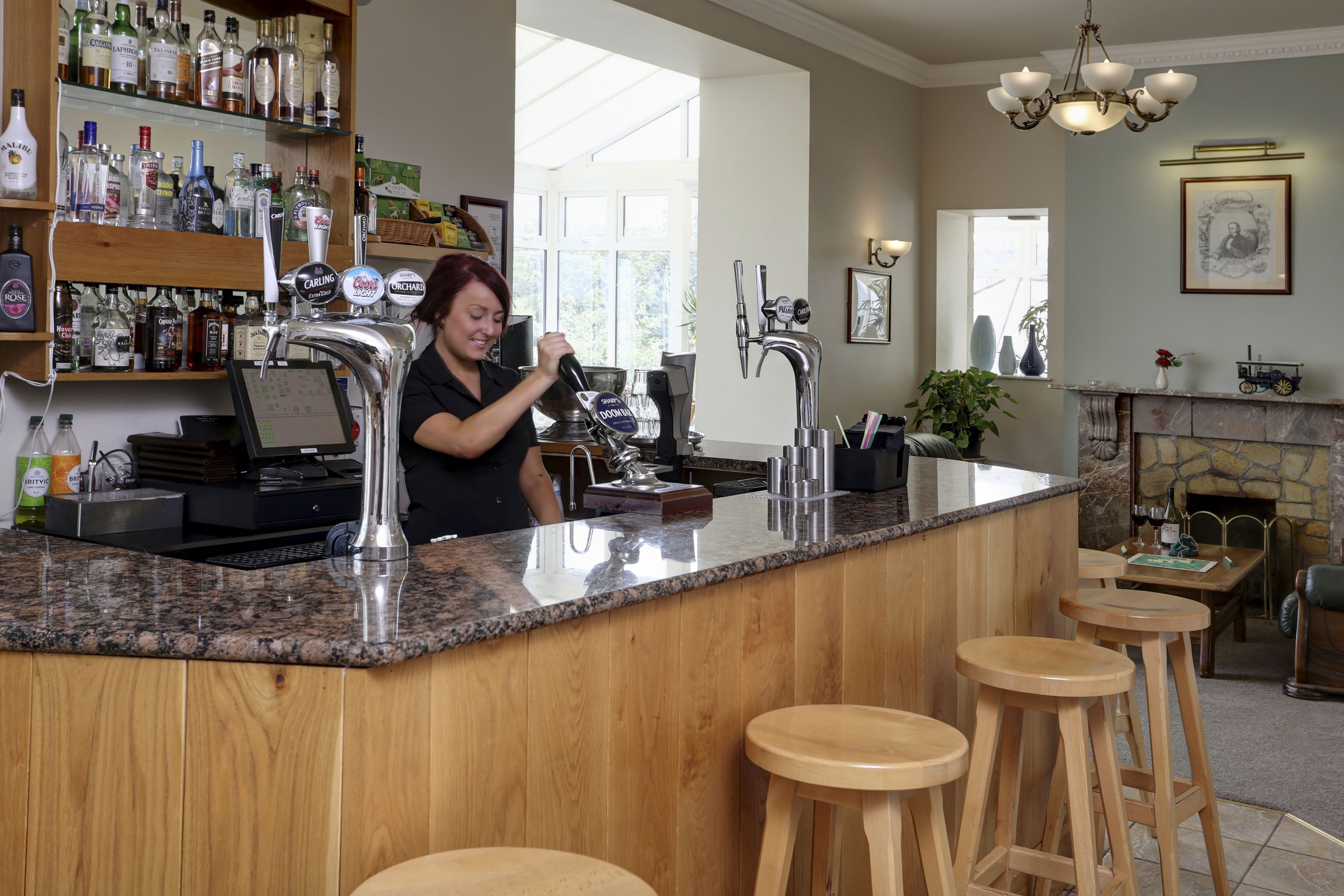 Uma área de bar com balcão de madeira, banquetas e diversas garrafas de bebidas nas prateleiras.