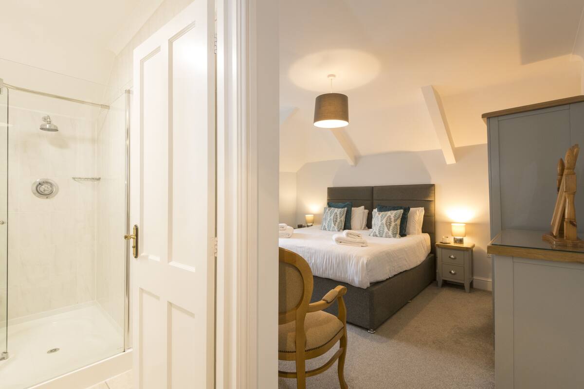 Habitación de hotel con cabina de ducha de vidrio, cama con cabecero, mesitas de noche con lámparas y una silla de madera.